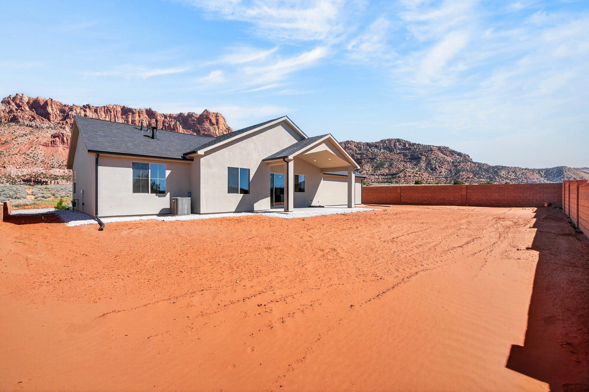 Contemporary stucco exterior with a covered patio, dark-framed windows, dark asphalt shingle roof, and a red rock mountain backdrop
