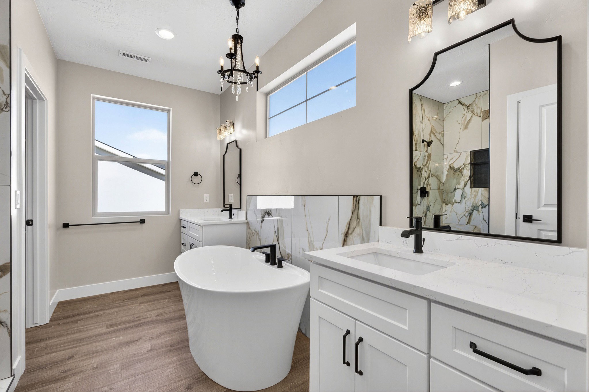 Spacious bathroom featuring a freestanding soaking tub, dual vanities with white countertops, wood-finish flooring, a contemporary black chandelier, and a walk-in shower with marble-look tile surround