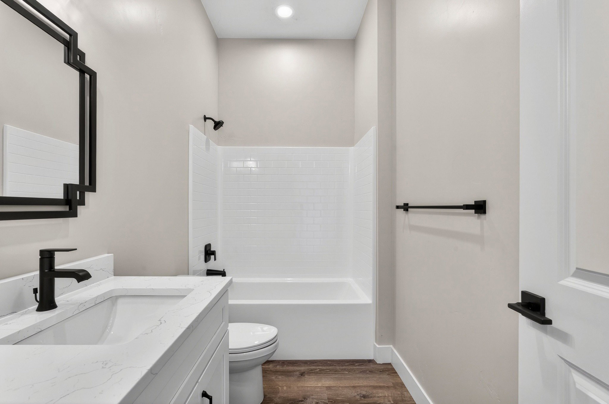 Bathroom featuring wood-finish flooring, a white vanity with an undermount sink and stone-finish countertop, a black framed mirror, a white bathtub with subway tile surround, and matte black fixtures
