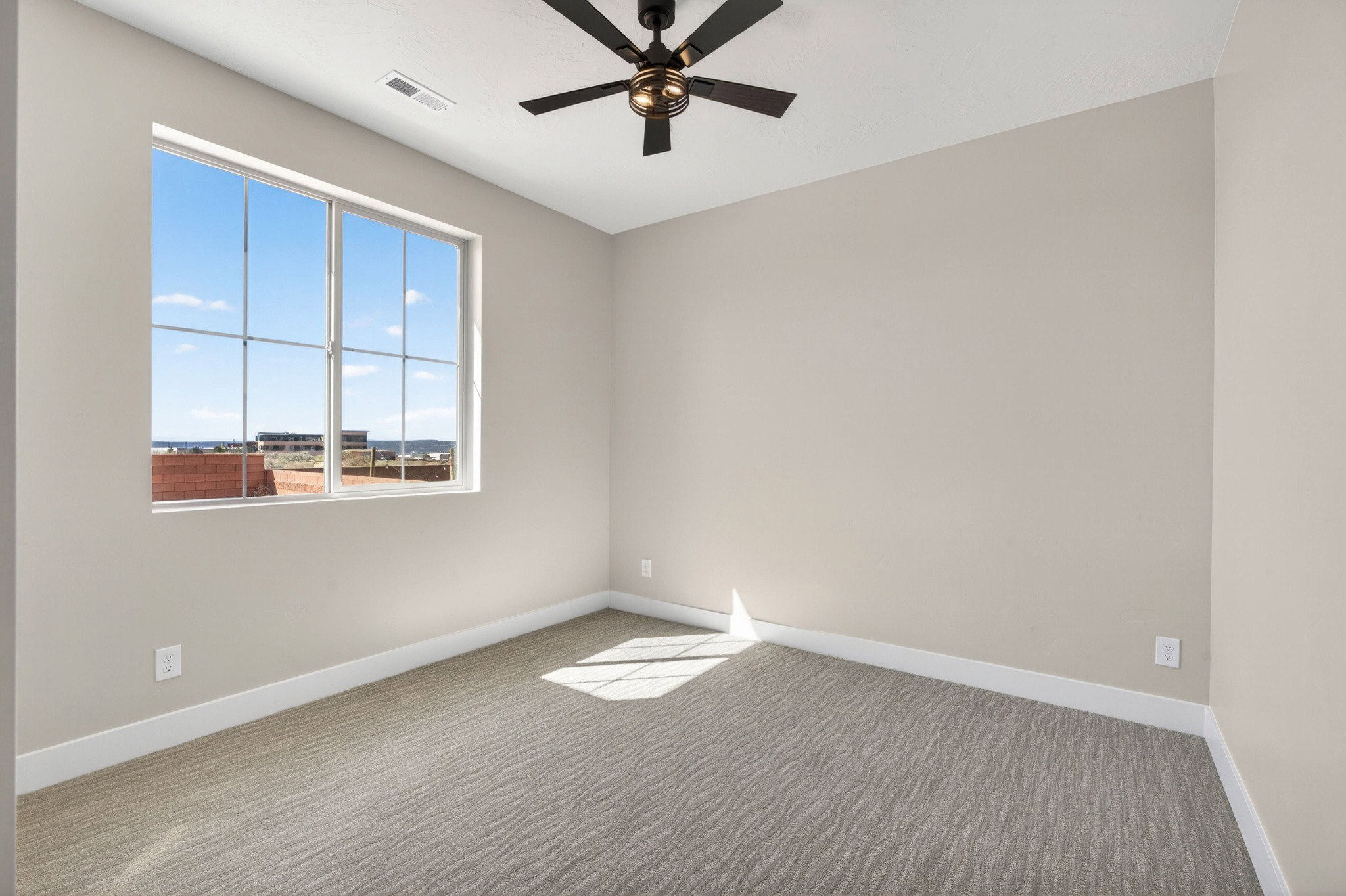 Room featuring a large window with gridded panes, a ceiling fan with dark blades and a bronze-toned fixture, light neutral-toned walls, and patterned carpeting