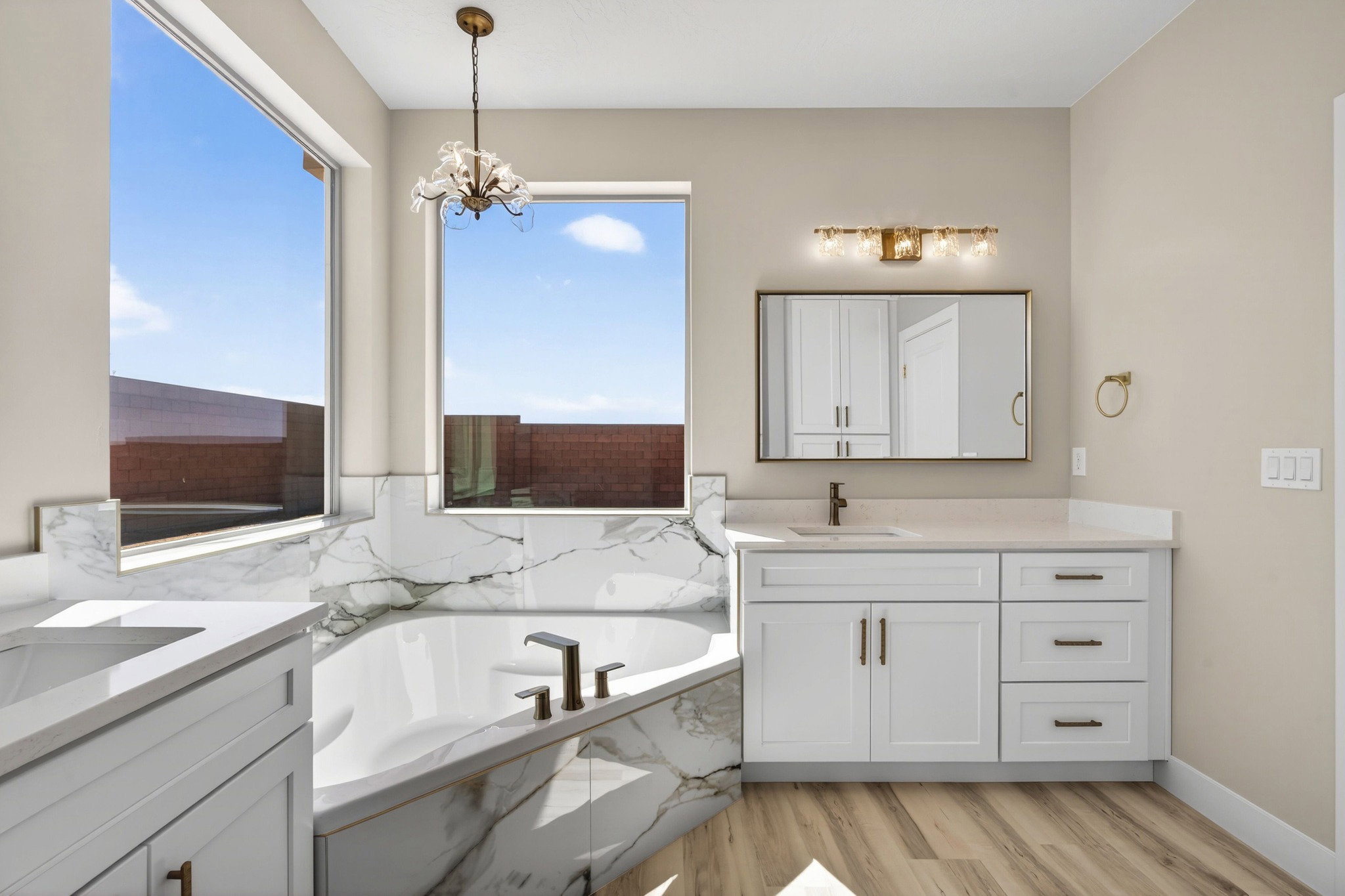 Soaking tub featuring a marble-patterned surround, dual windows, and an overhead chandelier