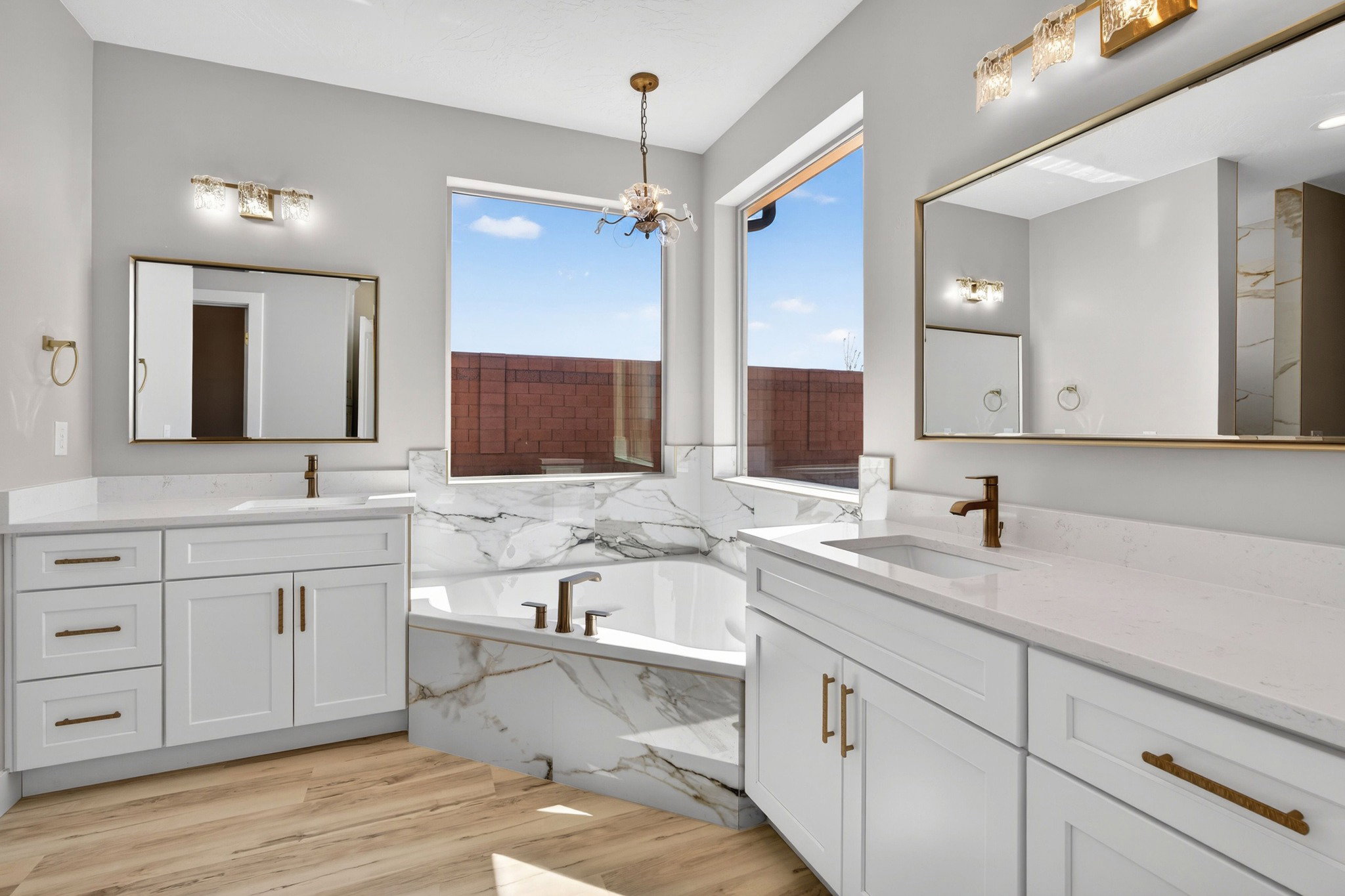 Bathroom featuring a large soaking tub with marble-look surround, dual white vanities with integrated sinks and quartz countertops, and wood-finish flooring