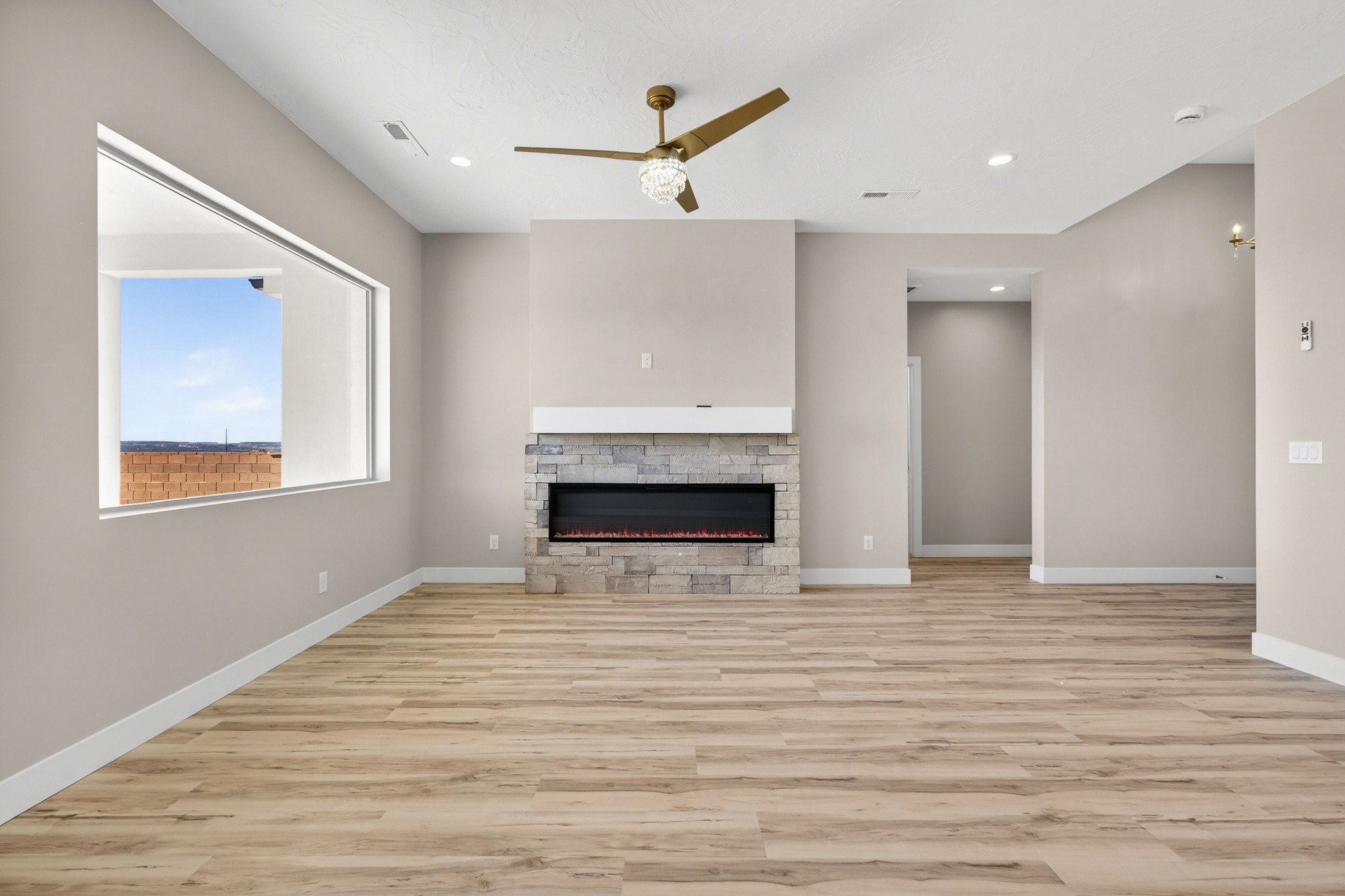 Spacious interior featuring wood-finish flooring, a contemporary fireplace with a stone surround and white mantel, a large horizontal window, a ceiling fan with integrated lighting, and recessed ceiling lights
