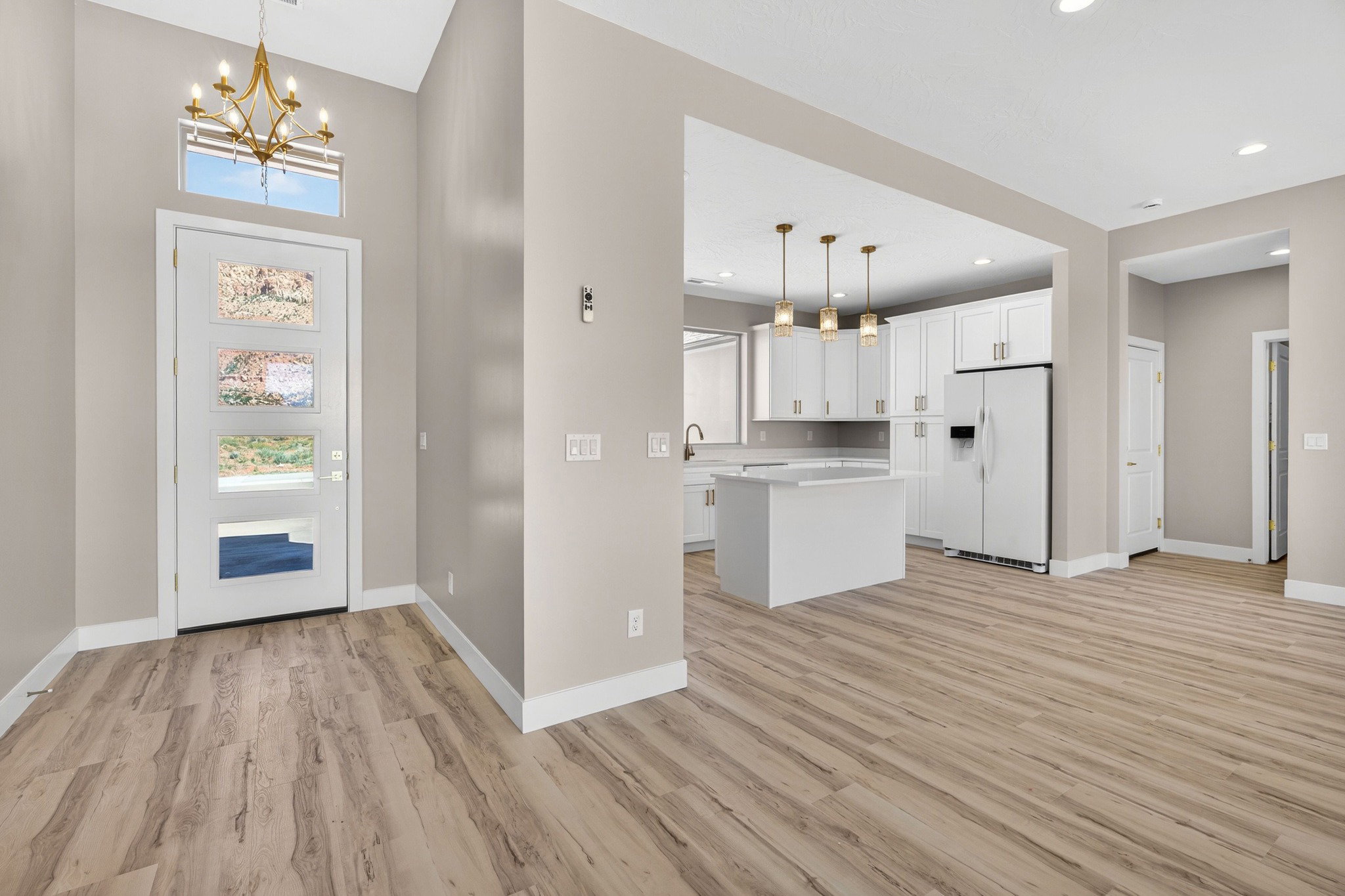 Open-concept living space featuring wood-finish flooring, a contemporary kitchen with white cabinetry and an island, a modern front entry door with obscured glass panels, a brass chandelier, and recessed lighting