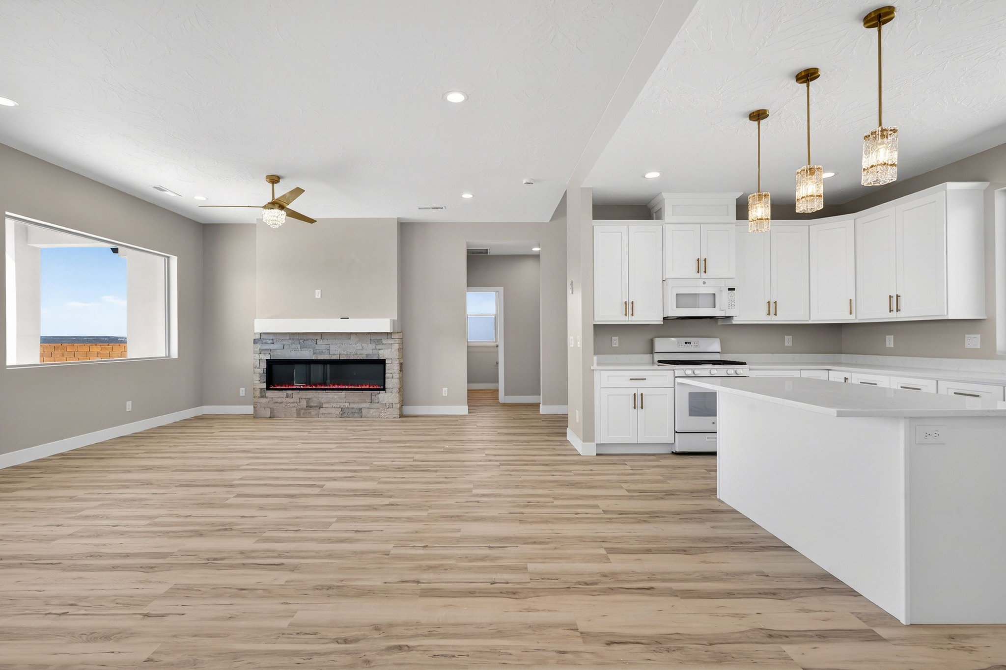 Open-concept living space featuring wood-finish flooring, a stone fireplace with a white mantel, and a kitchen with white cabinetry, white appliances, and a central island