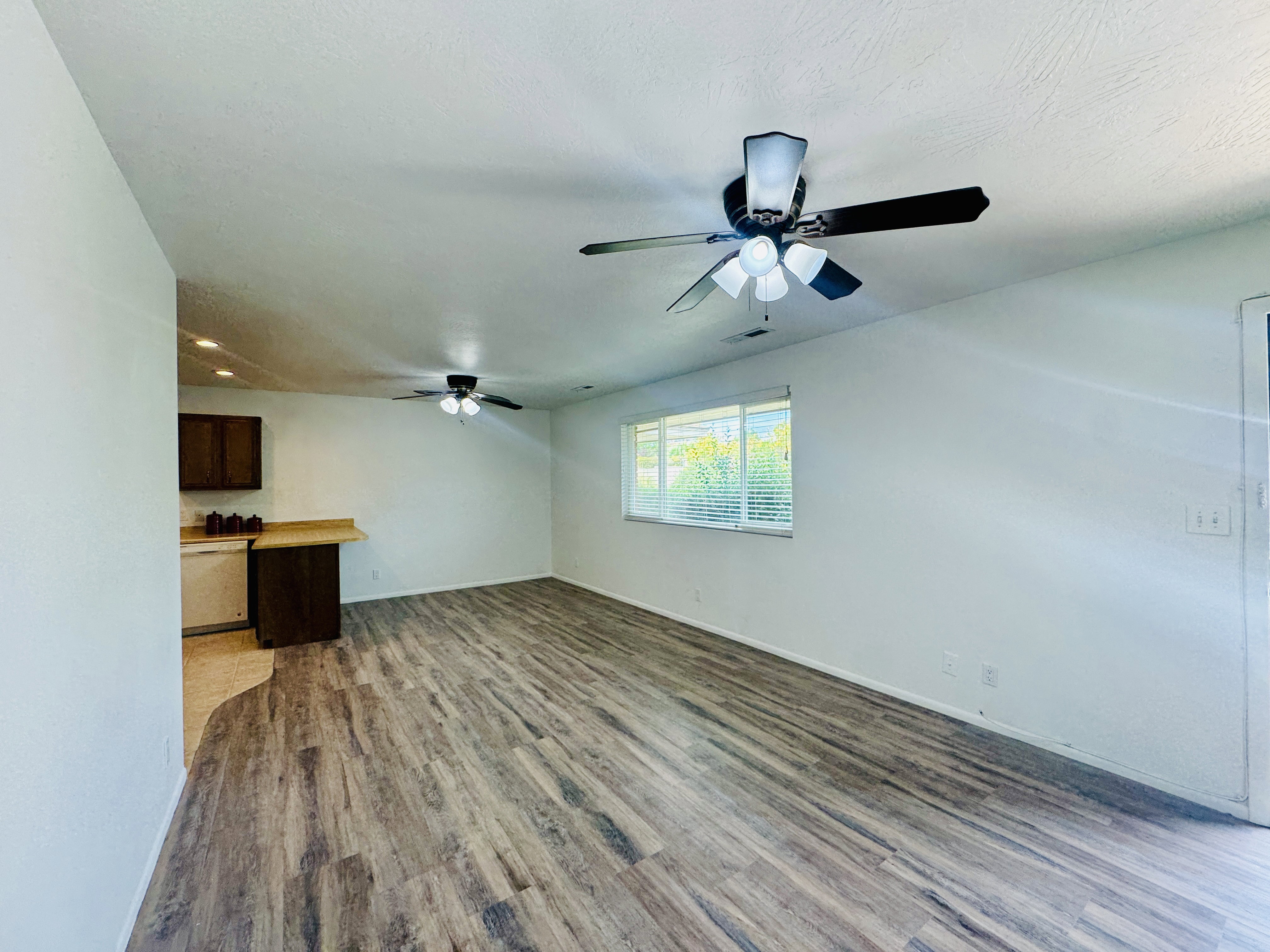 Spacious open-concept living area featuring wood-finish flooring, white walls, and a large window with blinds