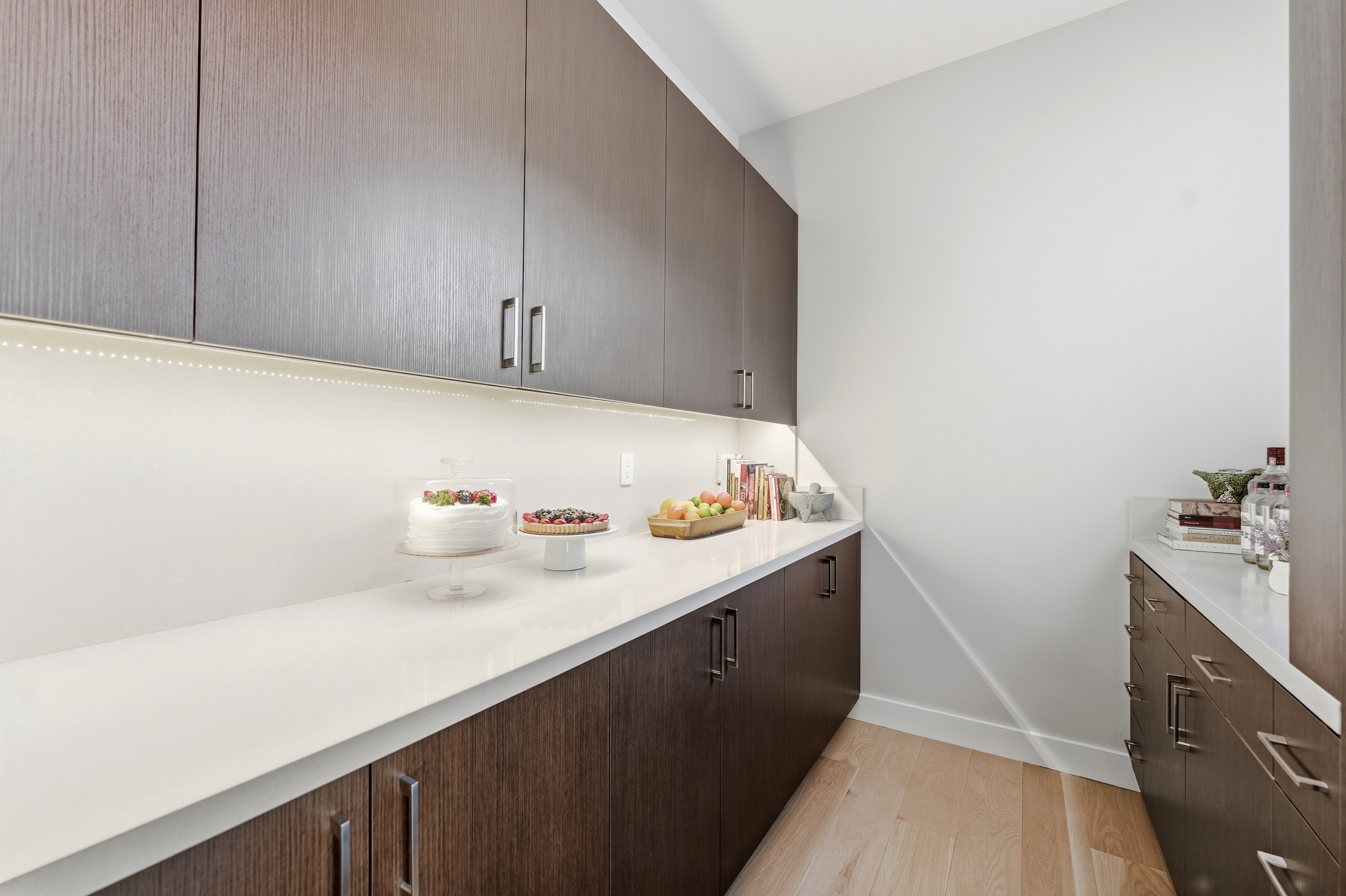 Modern pantry featuring wood-finish cabinetry, white countertops, under-cabinet lighting, and light wood-finish flooring