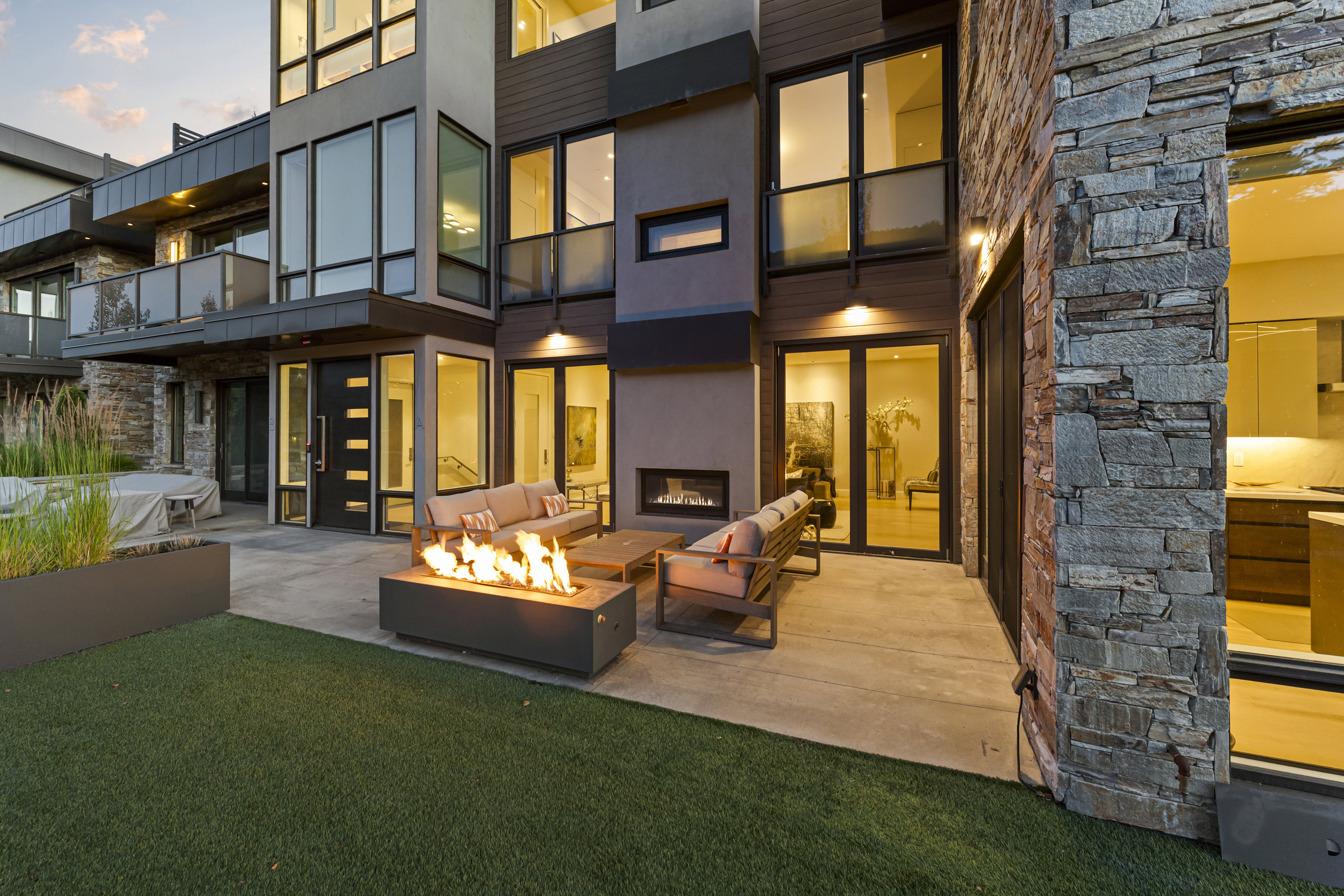 Modern exterior design featuring large format windows, a stone accent wall, a concrete patio with a built-in fire pit, a turf lawn, and a multi-level structure