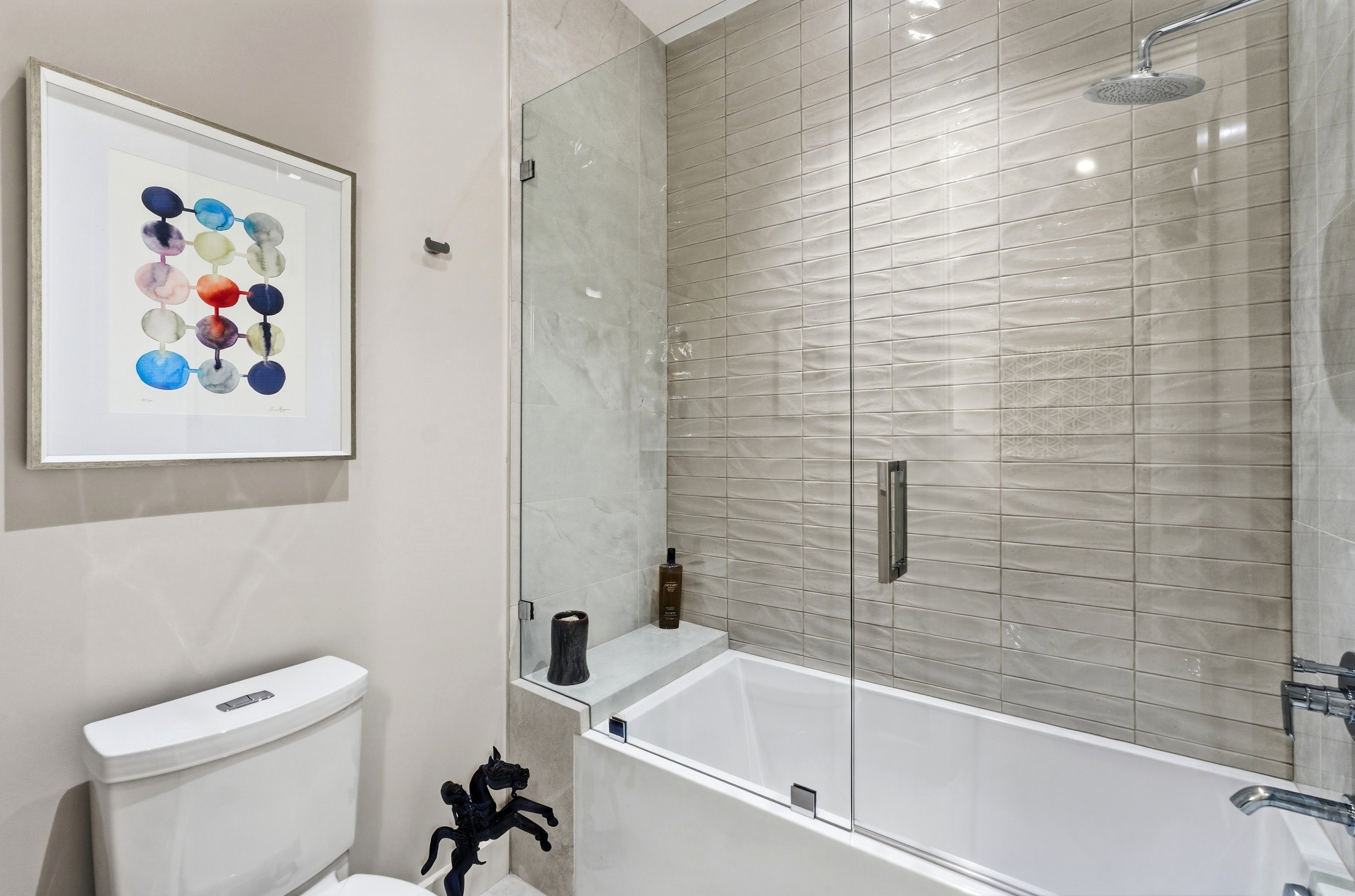 Contemporary bathroom featuring a white porcelain toilet, a tub with a glass enclosure, textured subway tile walls, a rain shower head, and a built-in tub shelf