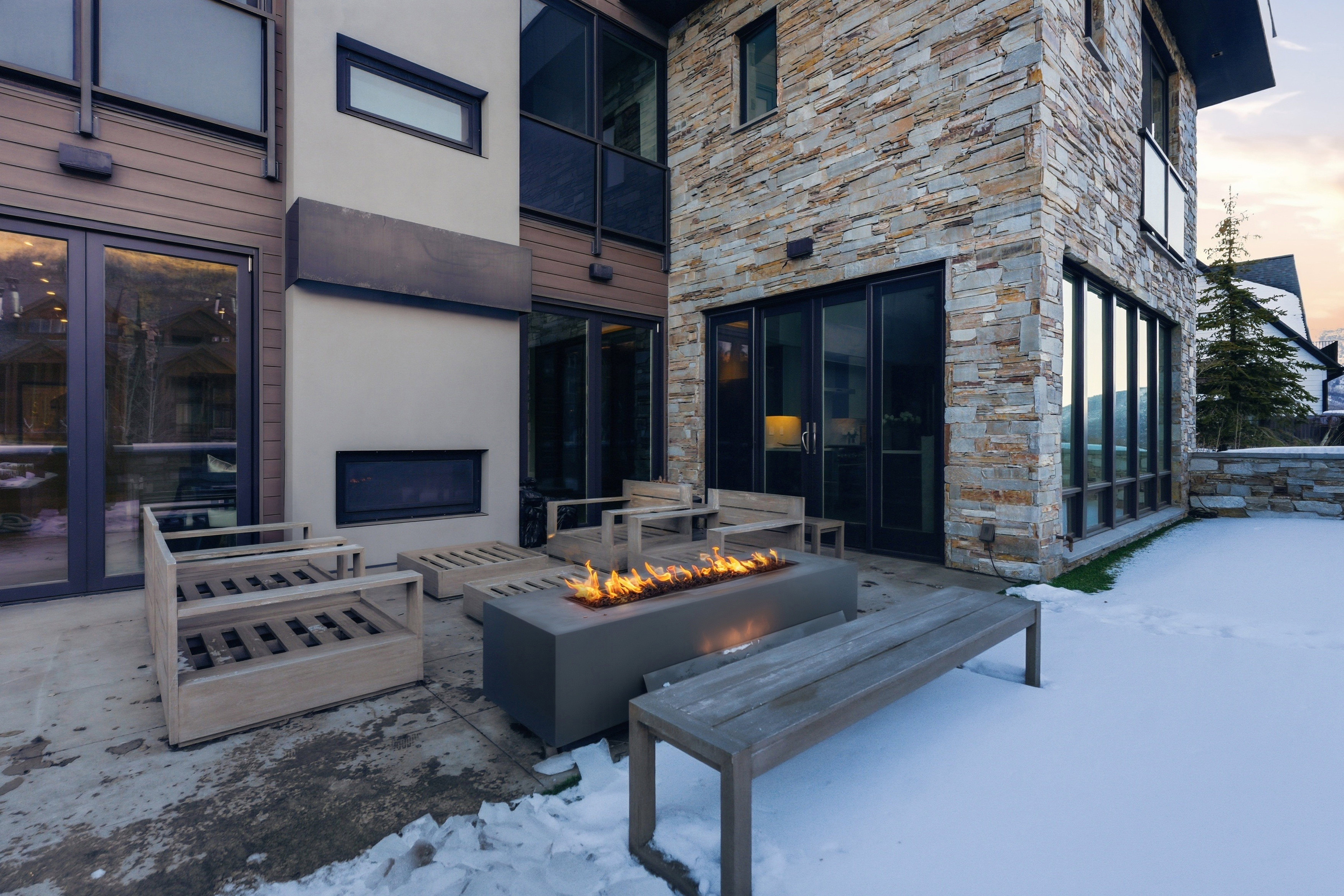 Contemporary stone and wood-paneled exterior featuring an outdoor fire pit, multiple large windows, and patio access