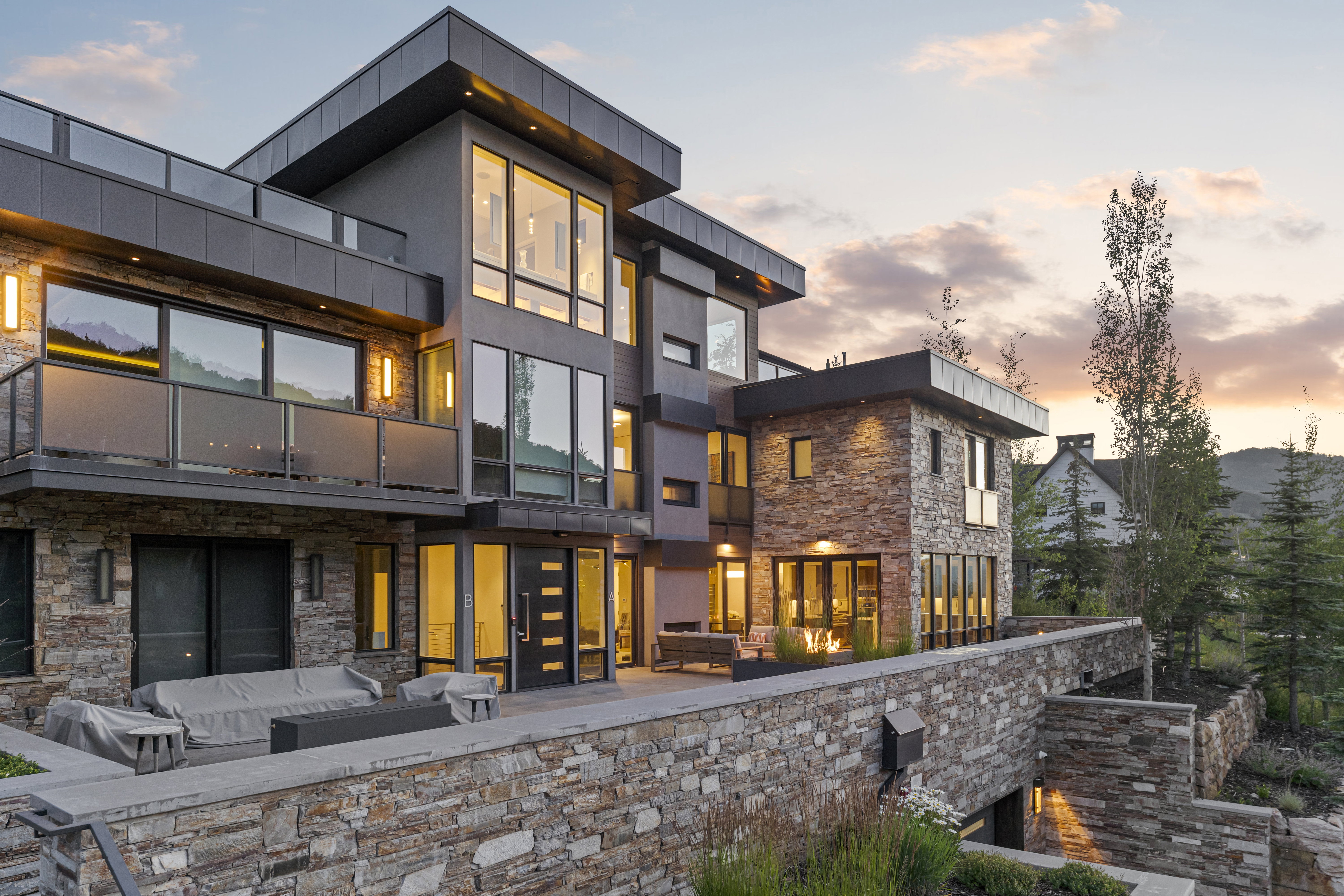 Contemporary facade featuring extensive stone cladding, large format windows, a flat roofline, multiple balconies with glass railings, and an integrated outdoor patio