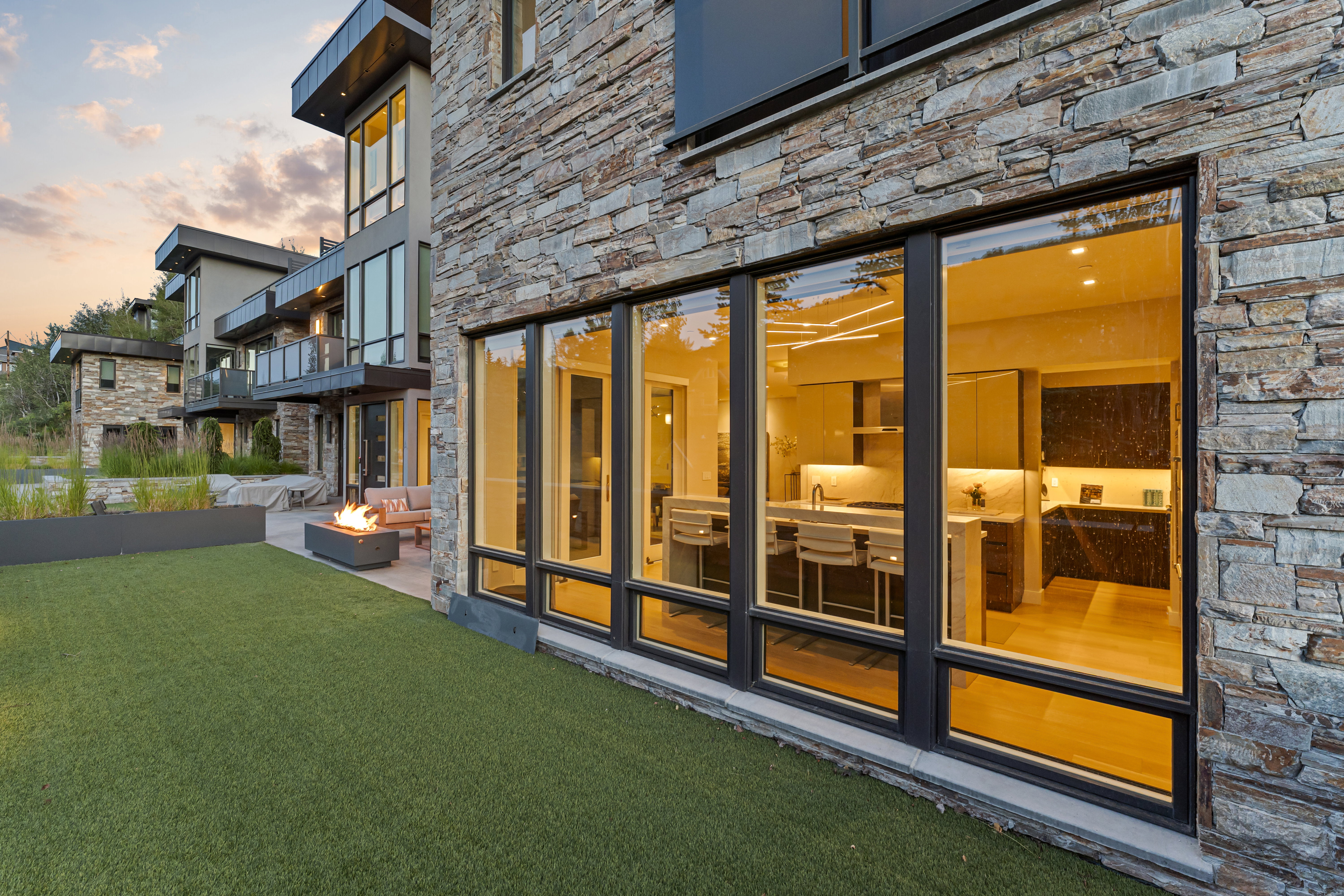 Stone veneer exterior with expansive black-framed windows revealing an illuminated kitchen interior