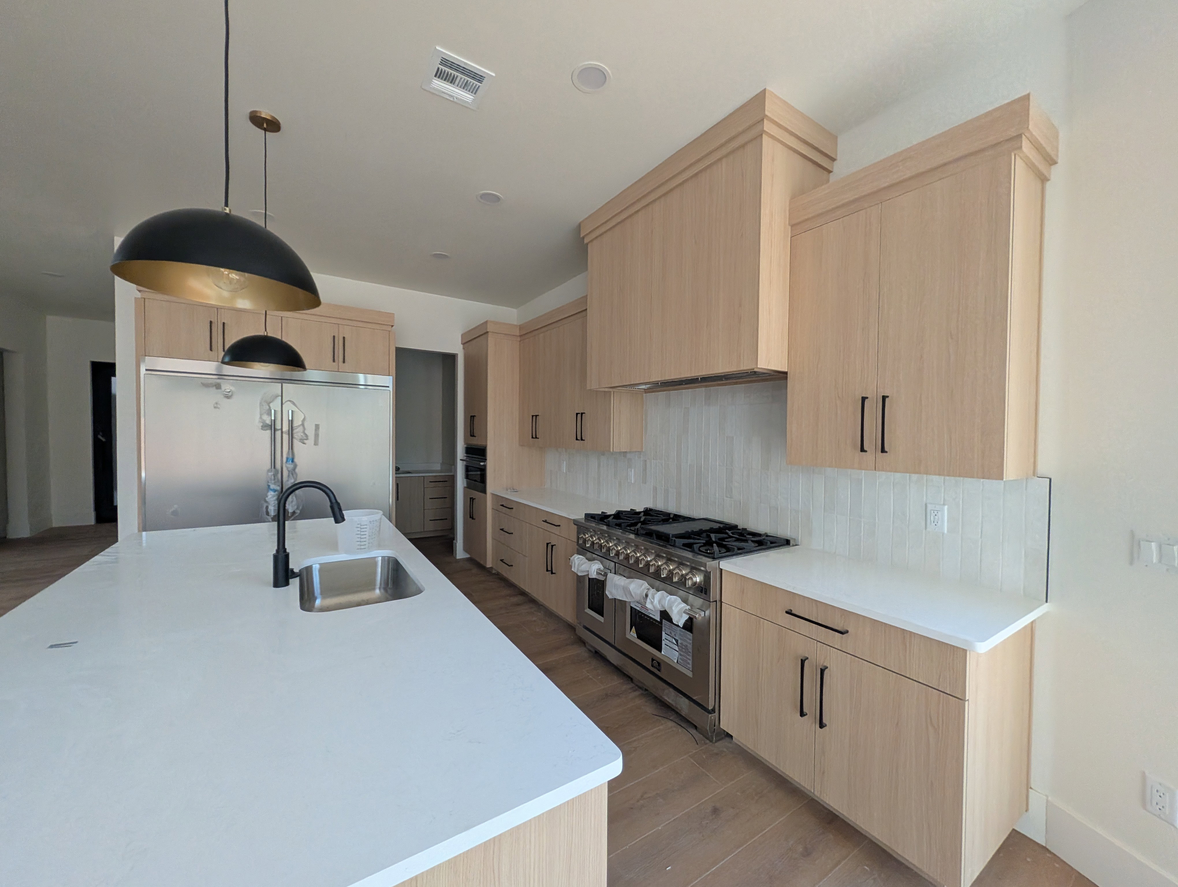 Gourmet kitchen featuring a substantial island with an undermount sink and matte black faucet, light wood-finish cabinetry, stainless steel appliances, a professional-grade range, and a full-height tile backsplash. Double wide fridge and gas range.