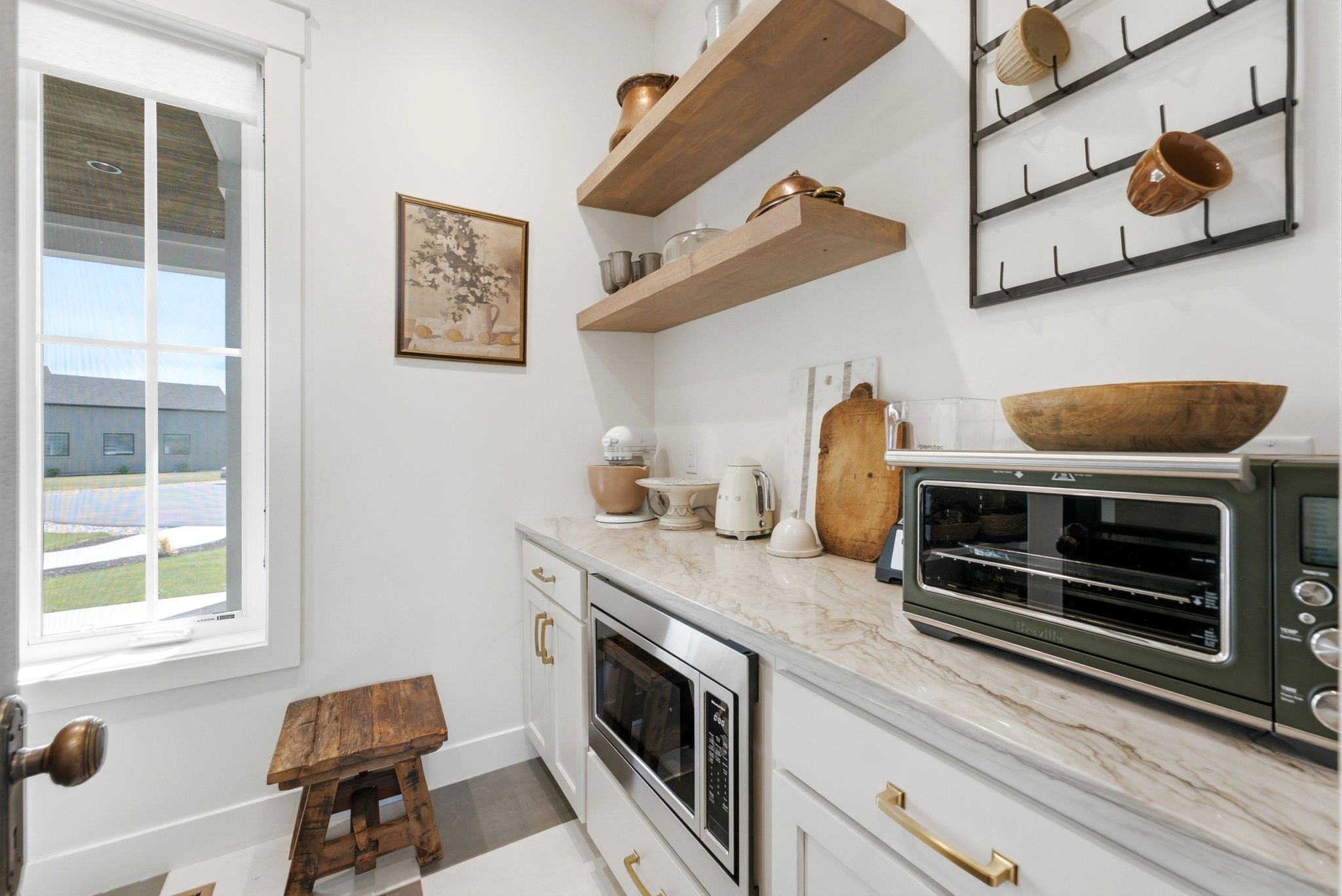 Built-in microwave, stone countertop, white cabinetry with brass hardware, open wood shelving, and a multi-pane window