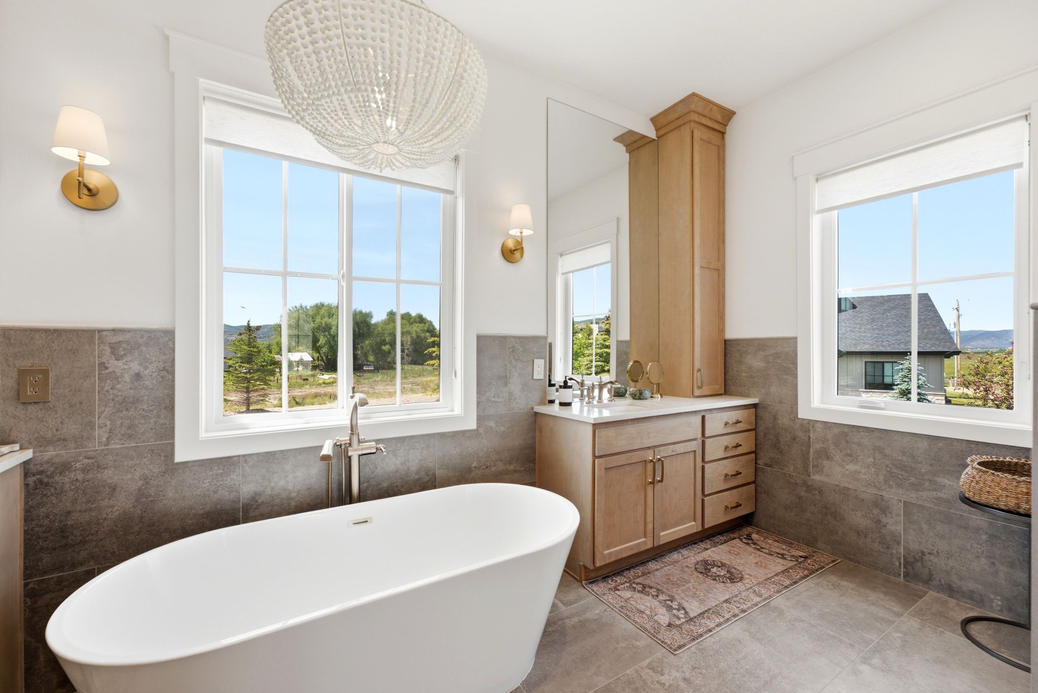 Bathroom featuring a freestanding oval tub with a floor-mounted faucet, dual windows, gray stone-look wall and floor tiling, and a double vanity with light wood cabinetry and a tall linen tower