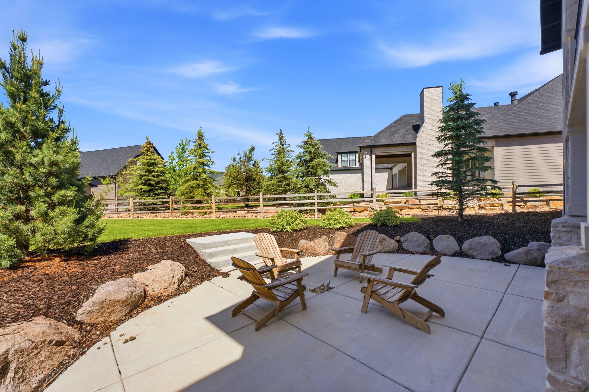 Spacious concrete patio with tiered landscaping, surrounded by a manicured lawn and mature evergreen trees