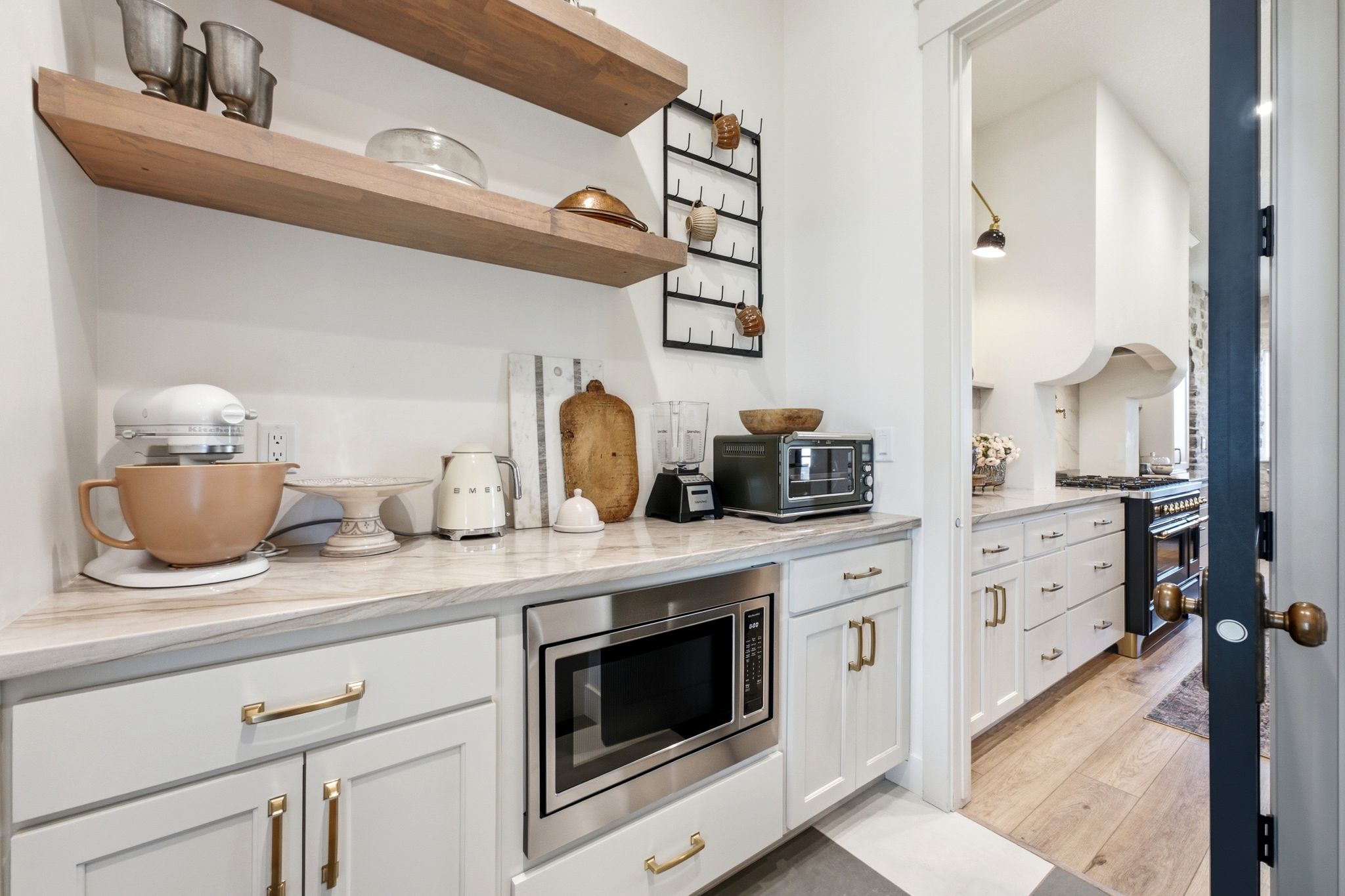 Dedicated butler's pantry featuring open wood shelving, light-toned countertops, white cabinetry with brass hardware, and a built-in microwave