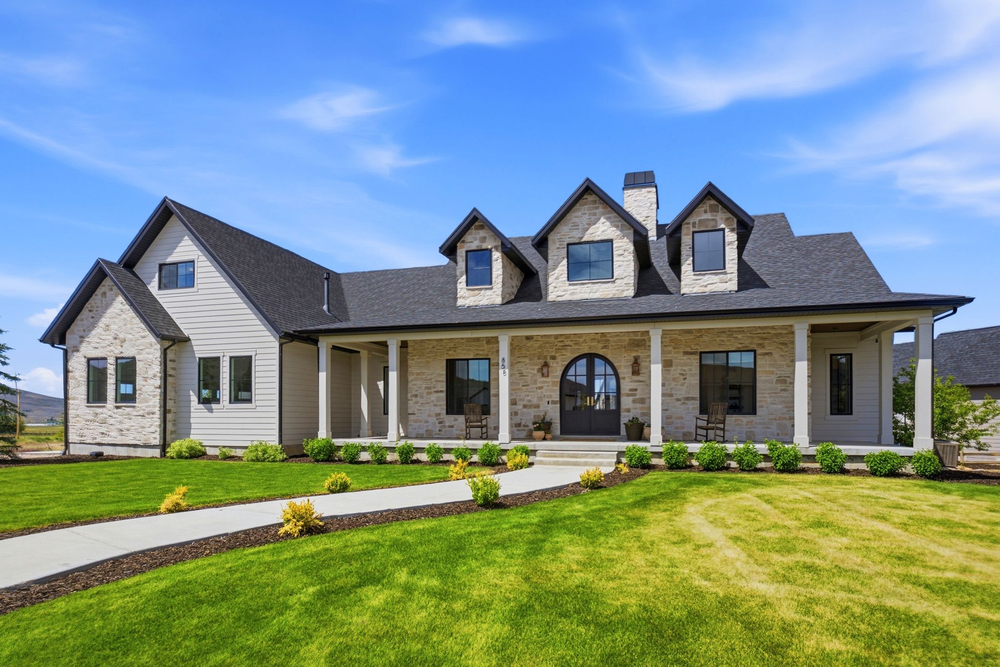 Stone and siding exterior with a covered front porch featuring multiple dormer windows, a prominent chimney, a dark shingled roof, and a paved walkway