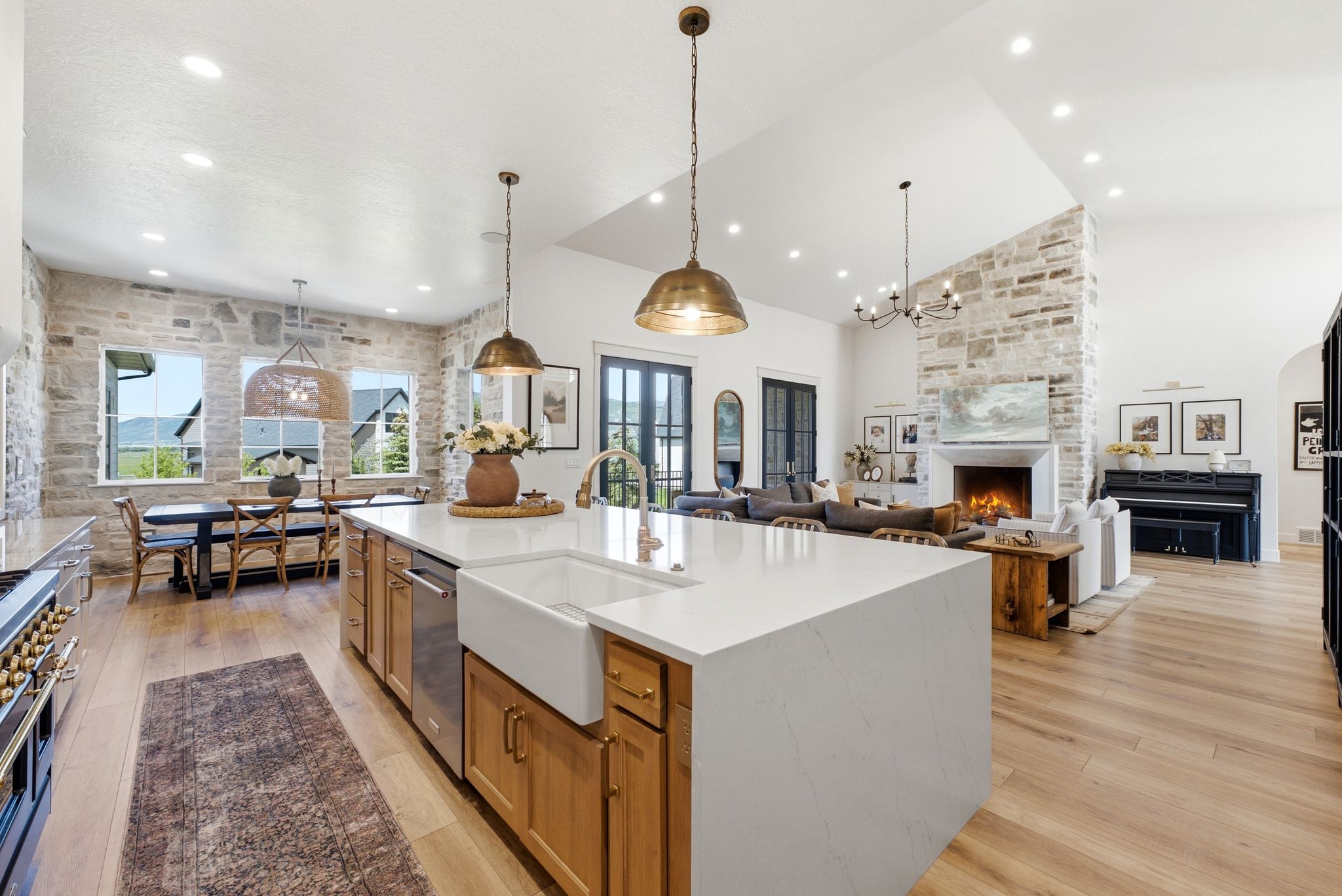 Spacious open-concept living area featuring an expansive kitchen island with a farmhouse sink, wood-finish flooring, a stone-accented fireplace, and vaulted ceilings