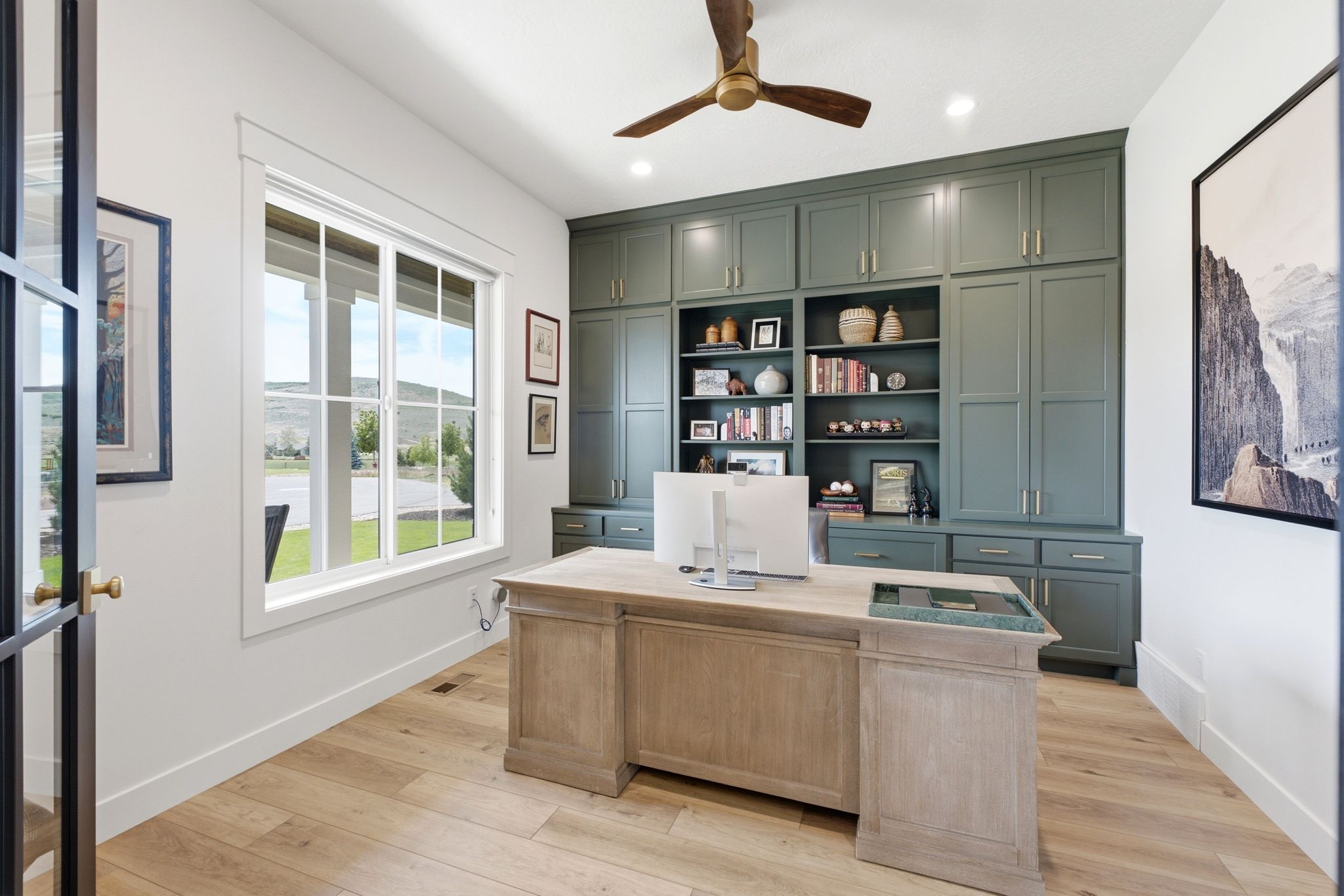 Dedicated office space featuring extensive built-in cabinetry, wood-finish flooring, a contemporary ceiling fan, and large window with exterior visibility