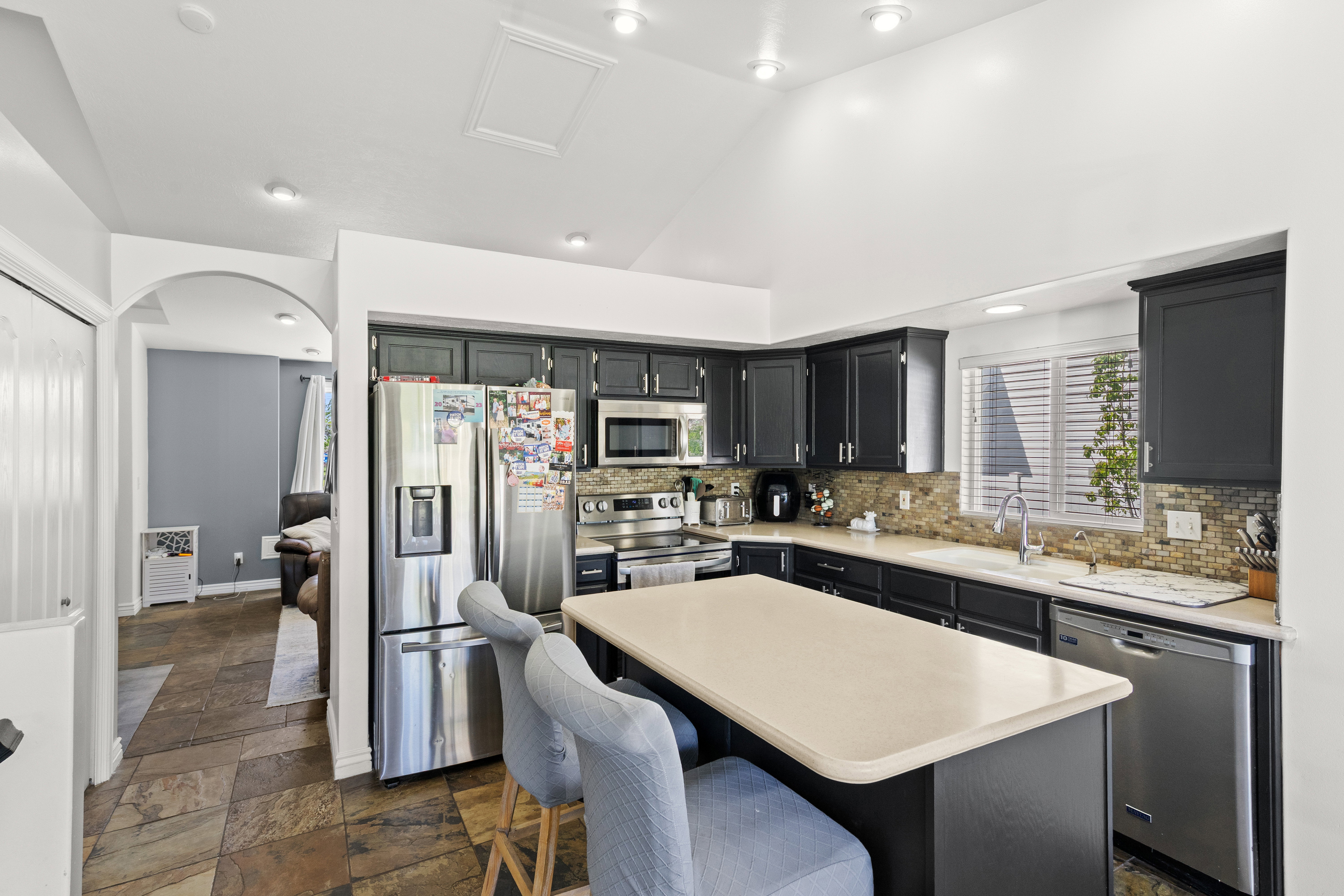 Kitchen featuring dark cabinetry, stainless steel appliances, a light-toned island with seating, a mosaic tile backsplash, and slate-look flooring