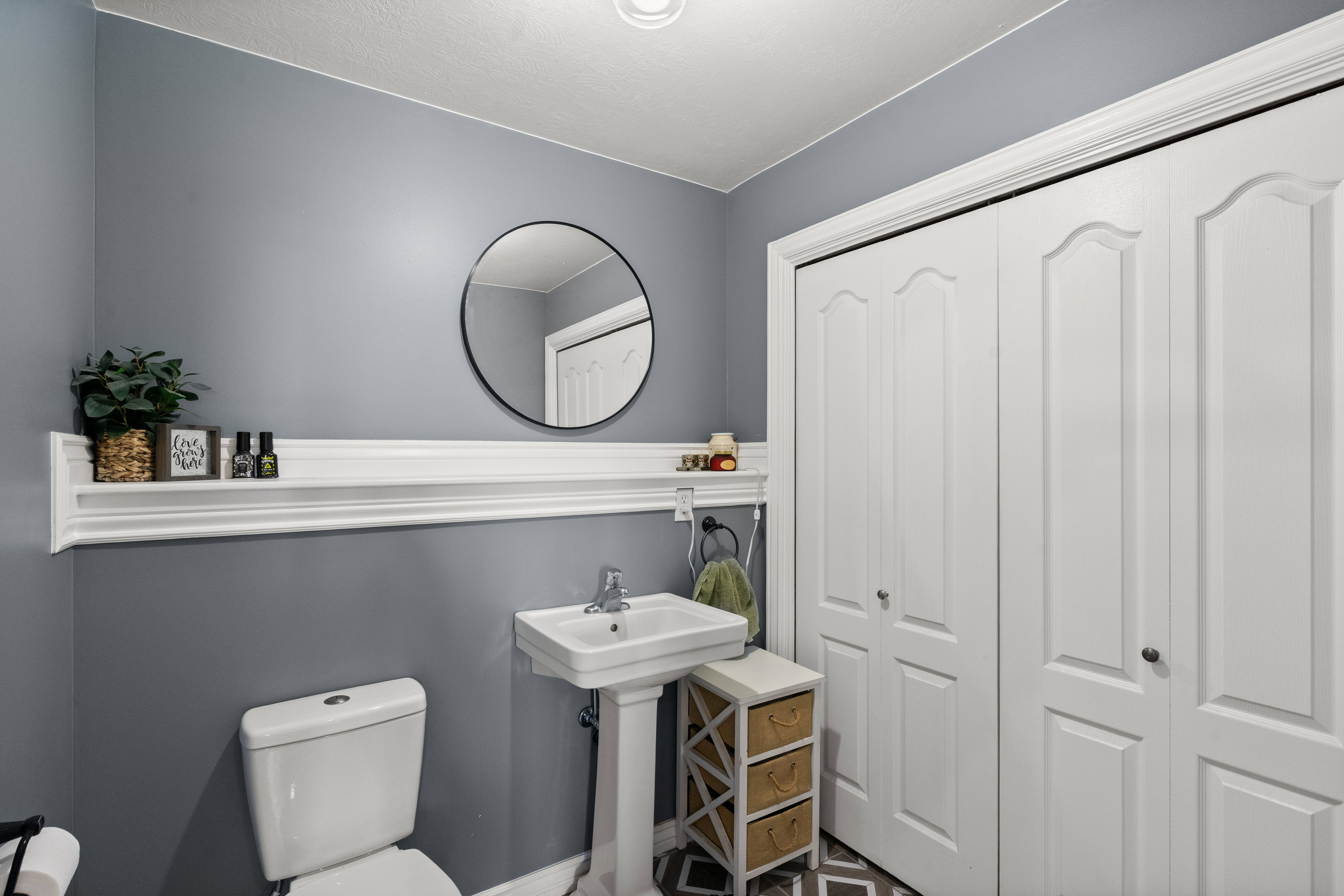Pedestal sink with chrome faucet, circular mirror, and wall-mounted shelving above the commode