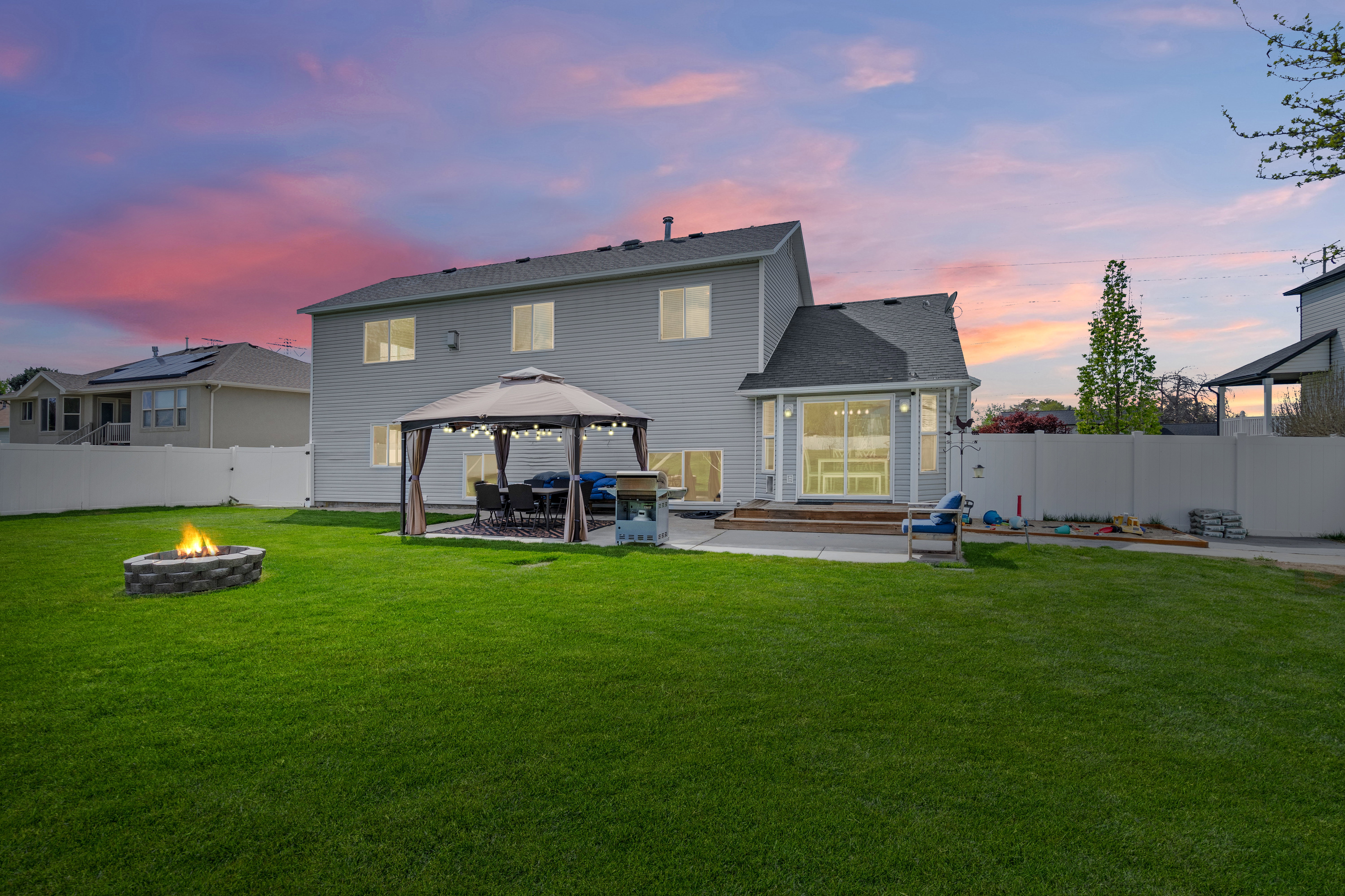 Expansive backyard featuring a large green lawn, a stone fire pit, and a patio area with a gazebo