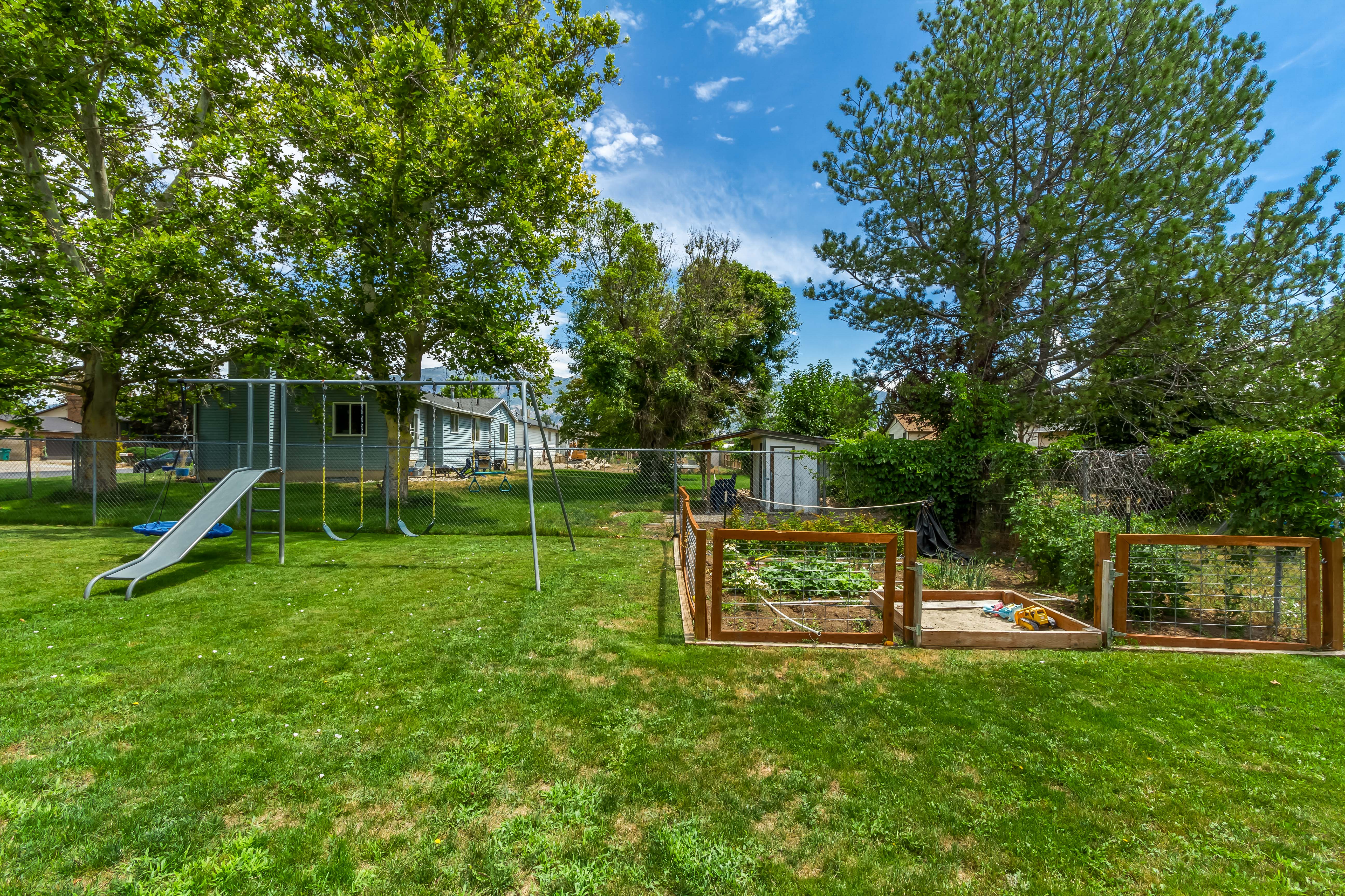 Expansive green lawn featuring a dedicated garden area with wooden raised beds and fencing, a swingset with slide, and mature trees providing shade