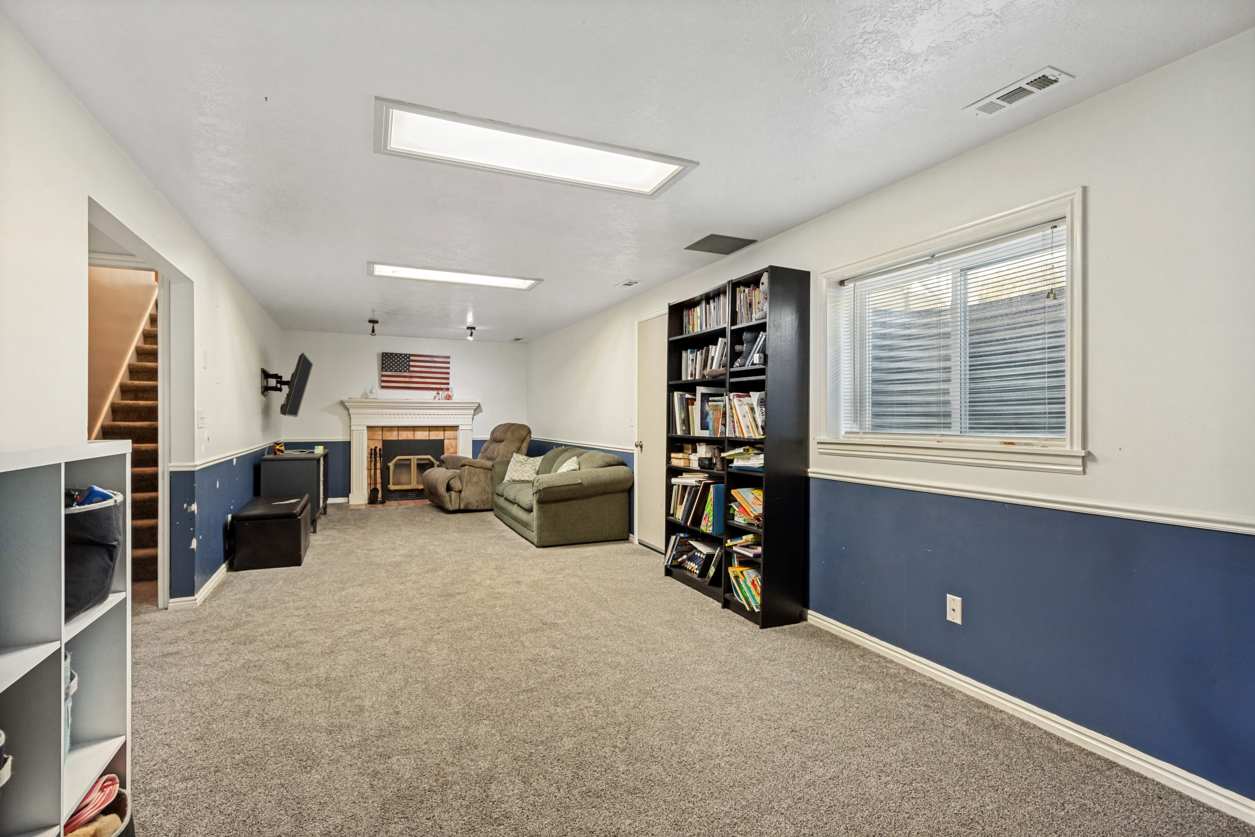 Spacious carpeted room featuring a white mantel fireplace, multiple ceiling light fixtures, a staircase with carpeted treads, and a built-in window
