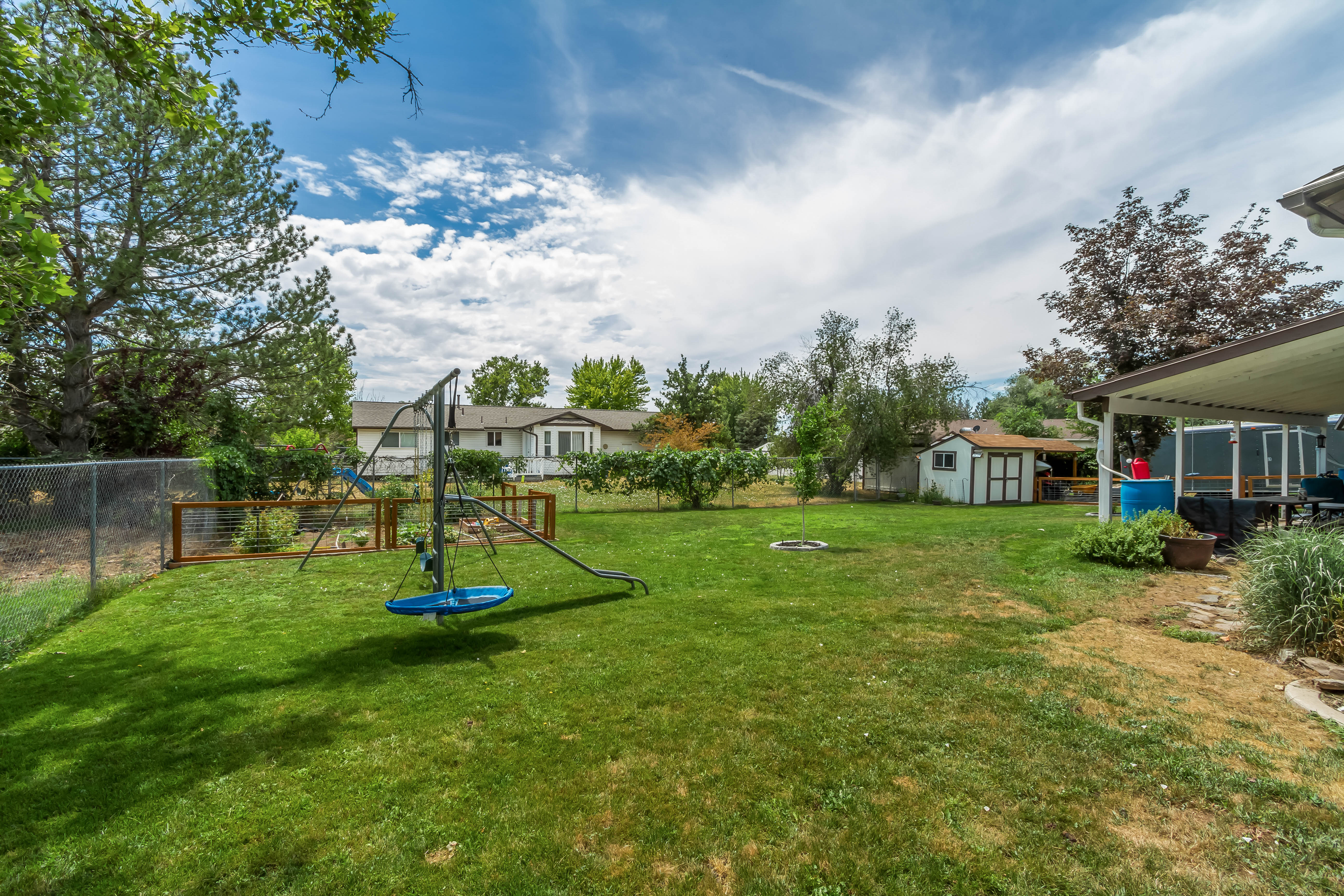 Expansive backyard featuring a large lawn area, chain-link fencing, a covered patio structure, mature trees, and a storage shed