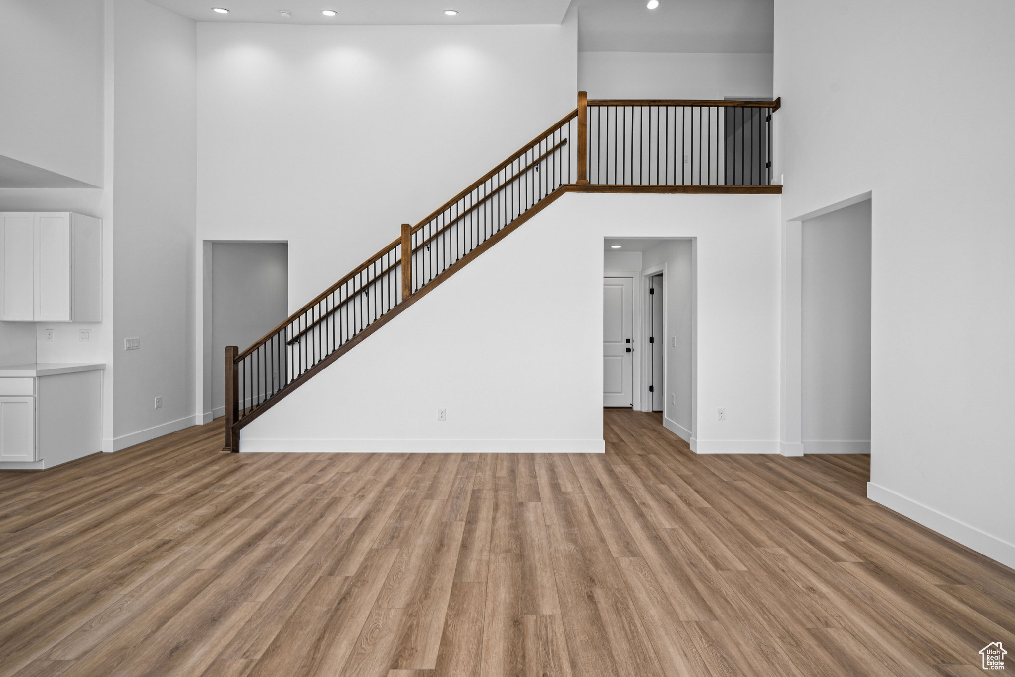 Open-concept living area featuring high ceilings, wood-finish flooring, a modern staircase with dark wood handrails and black balusters, recessed lighting, and crisp white walls
