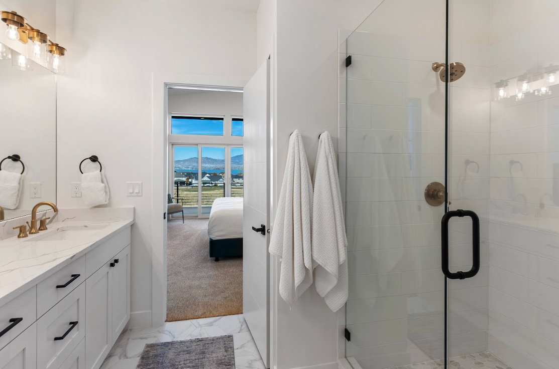 Bathroom featuring a spacious walk-in shower with clear glass enclosure and bronze fixtures, a vanity with a light-toned countertop and gold-toned faucet, and crisp white cabinetry with black hardware