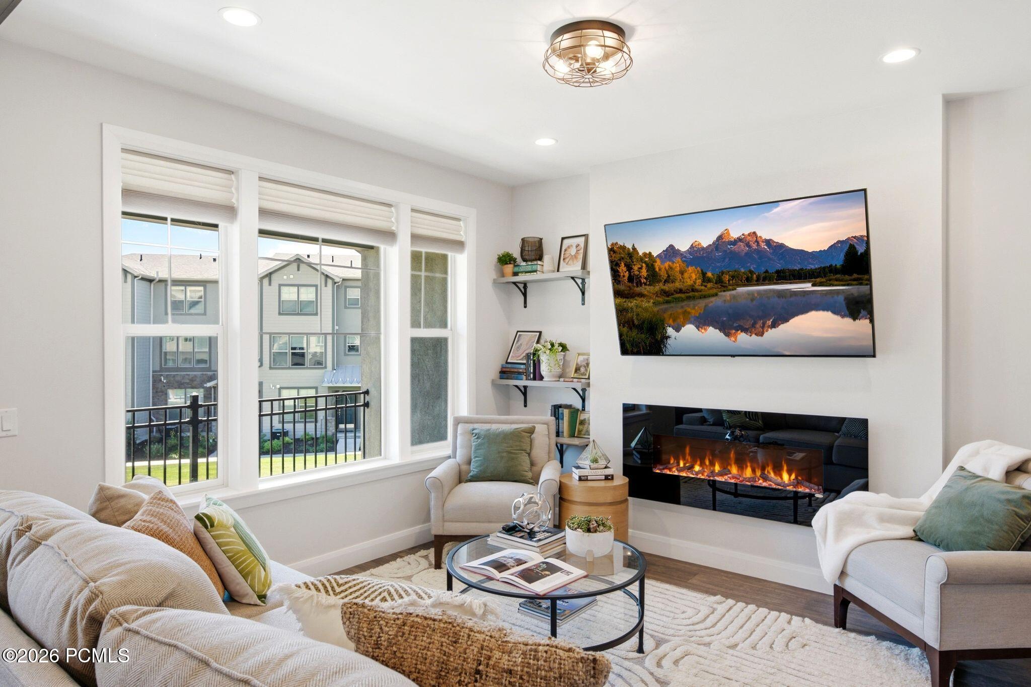 Living area featuring a sleek wall-mounted electric fireplace, multiple recessed ceiling lights, a contemporary semi-flush mount light fixture, and wood-finish flooring