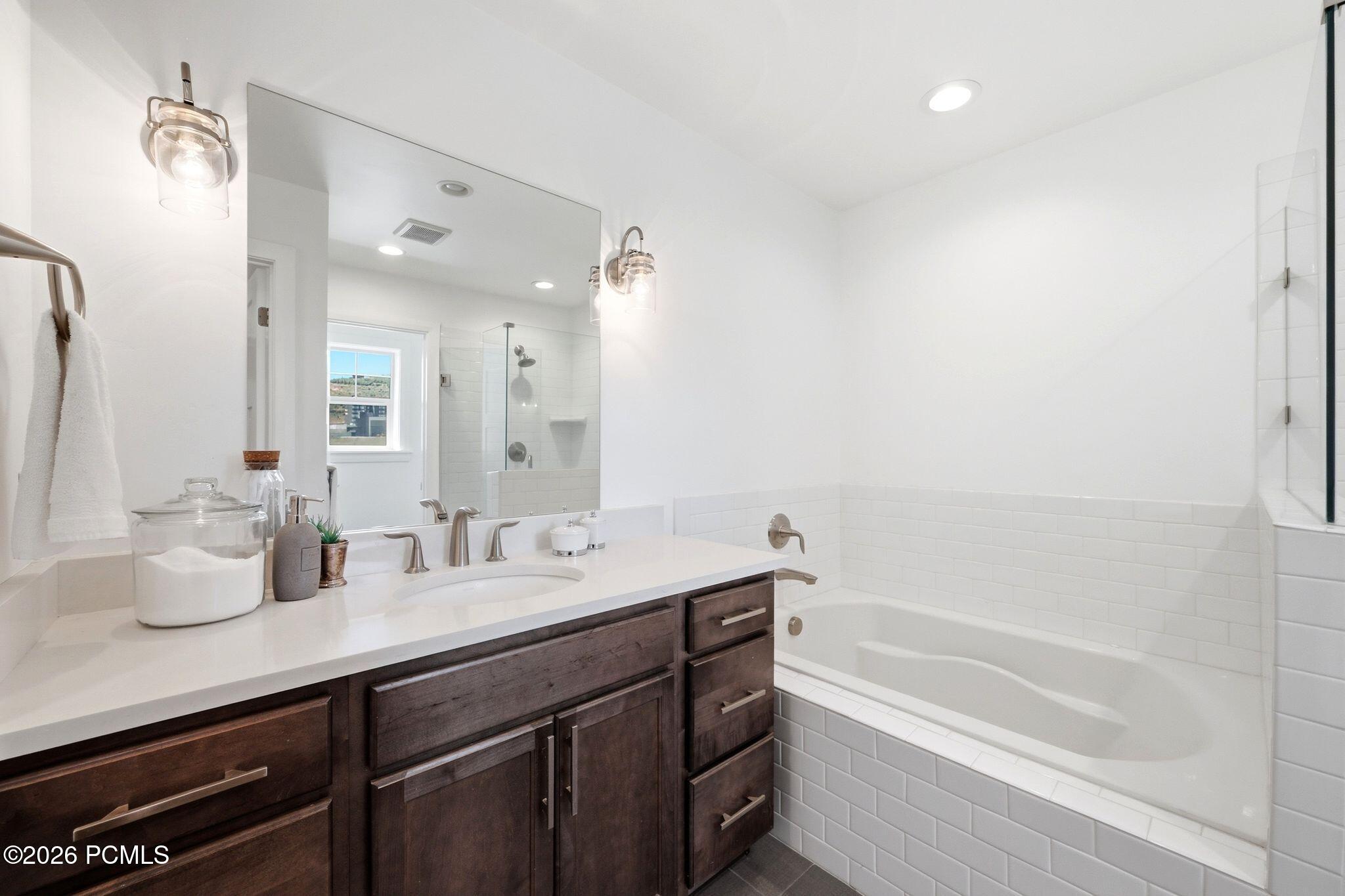 Bathroom featuring a deep soaking tub, subway tile surround, dark wood-finish vanity, and a large mirror with dual sconce lighting