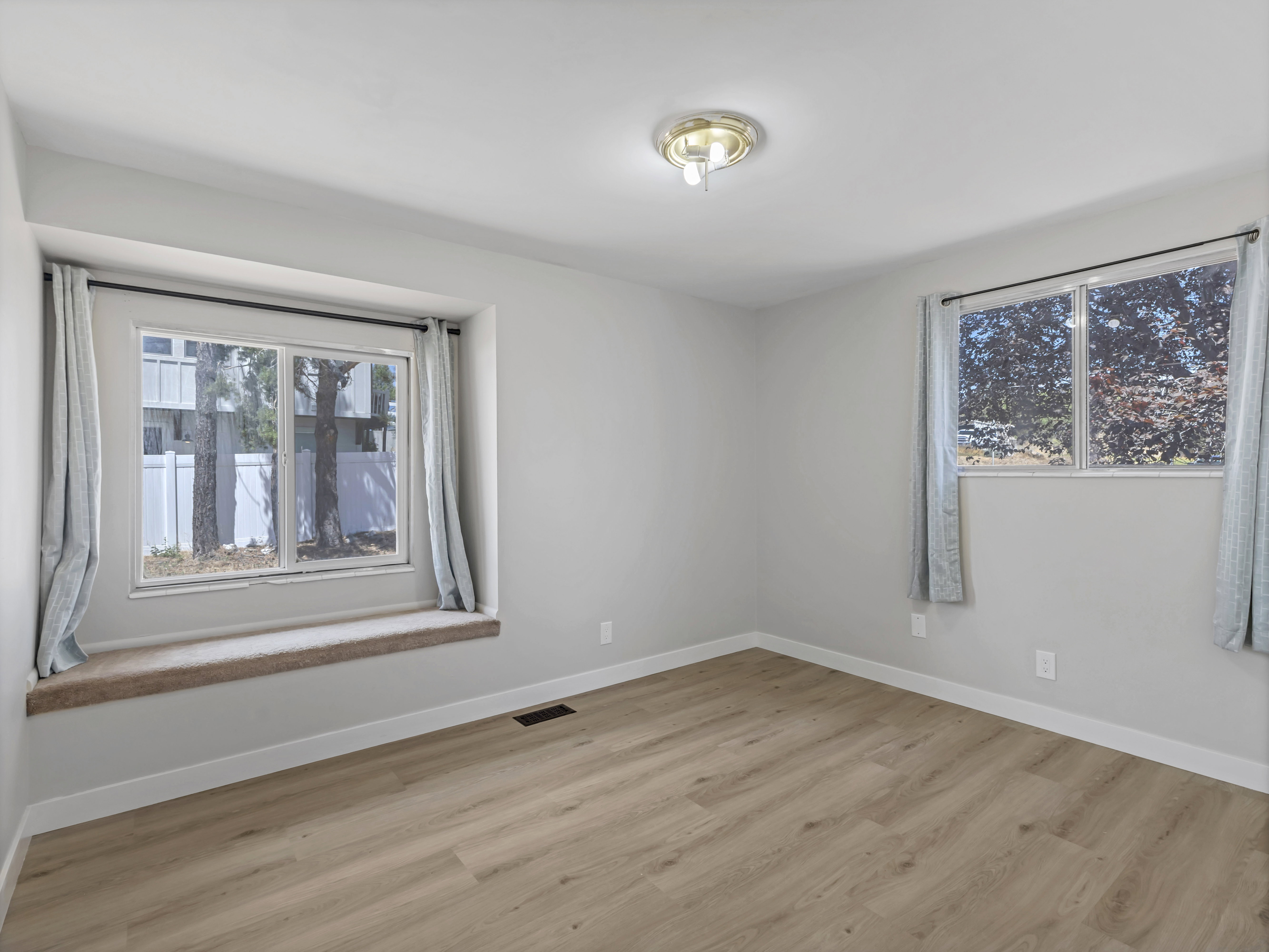 Bright interior space featuring light wood-finish flooring, a window seat with carpeted top, two windows with drapery rods, and a flush-mount ceiling light fixture