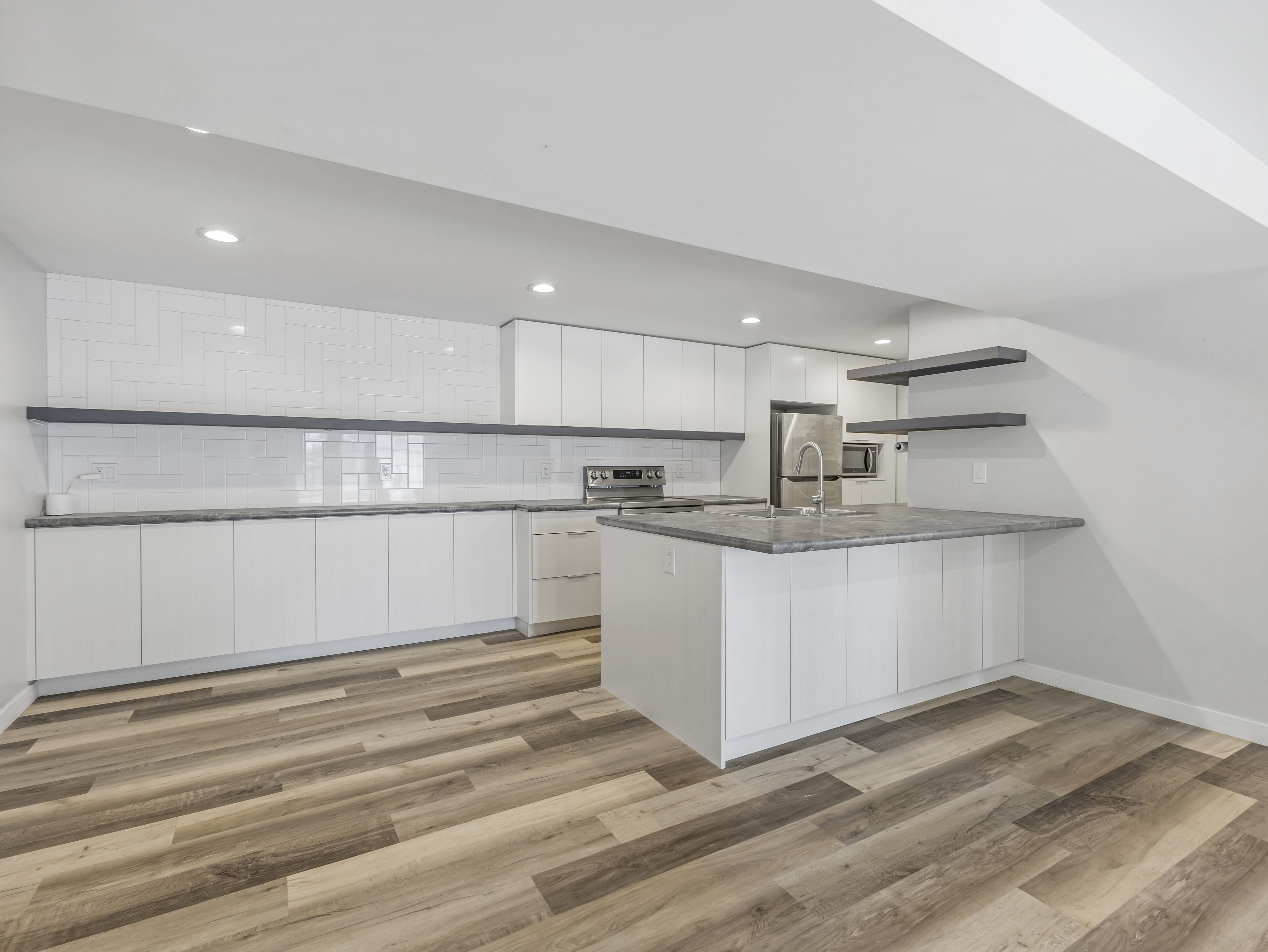 Modern kitchen featuring white flat-panel cabinetry, a central island with an undermount sink, stainless steel appliances, grey stone countertops, and wood-finish flooring