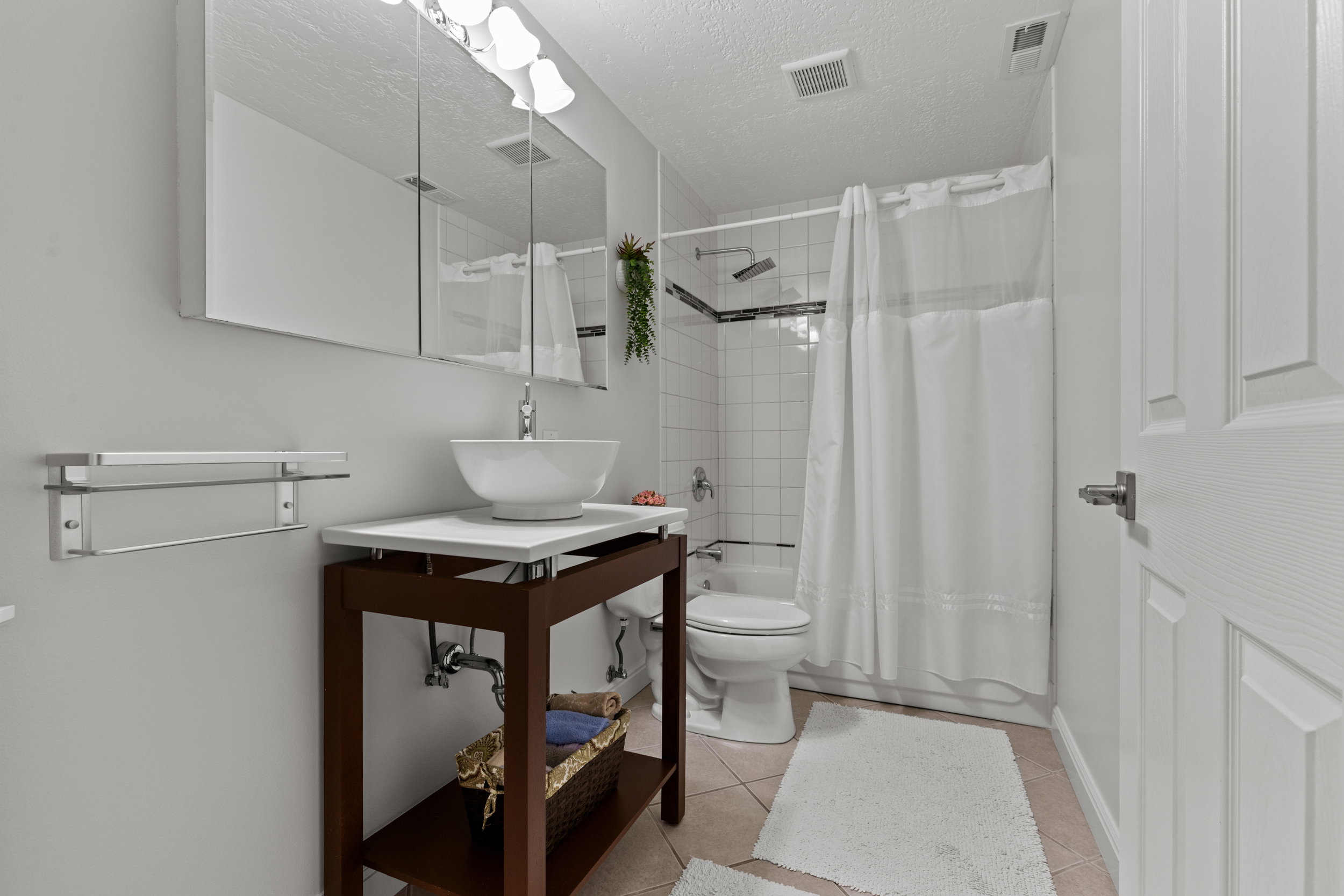 Contemporary bathroom featuring a vessel sink on a dark wood-finish vanity, a wall-mounted mirrored medicine cabinet, a tiled shower with a black accent border, a white paneled door, and neutral-toned floor tiling