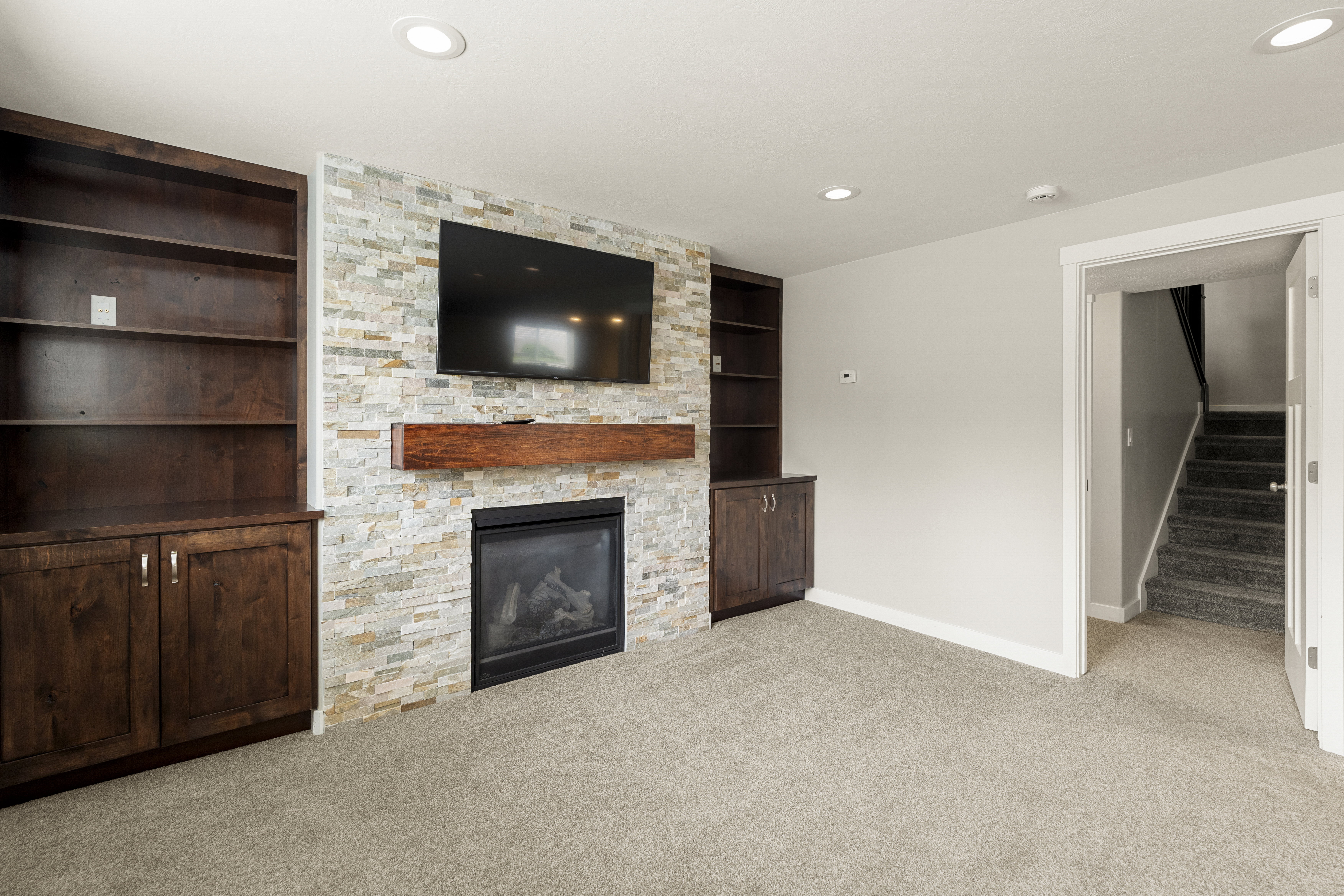 Carpeted room featuring a stone-clad fireplace with a wood mantle, recessed lighting, and built-in wood cabinetry with shelving