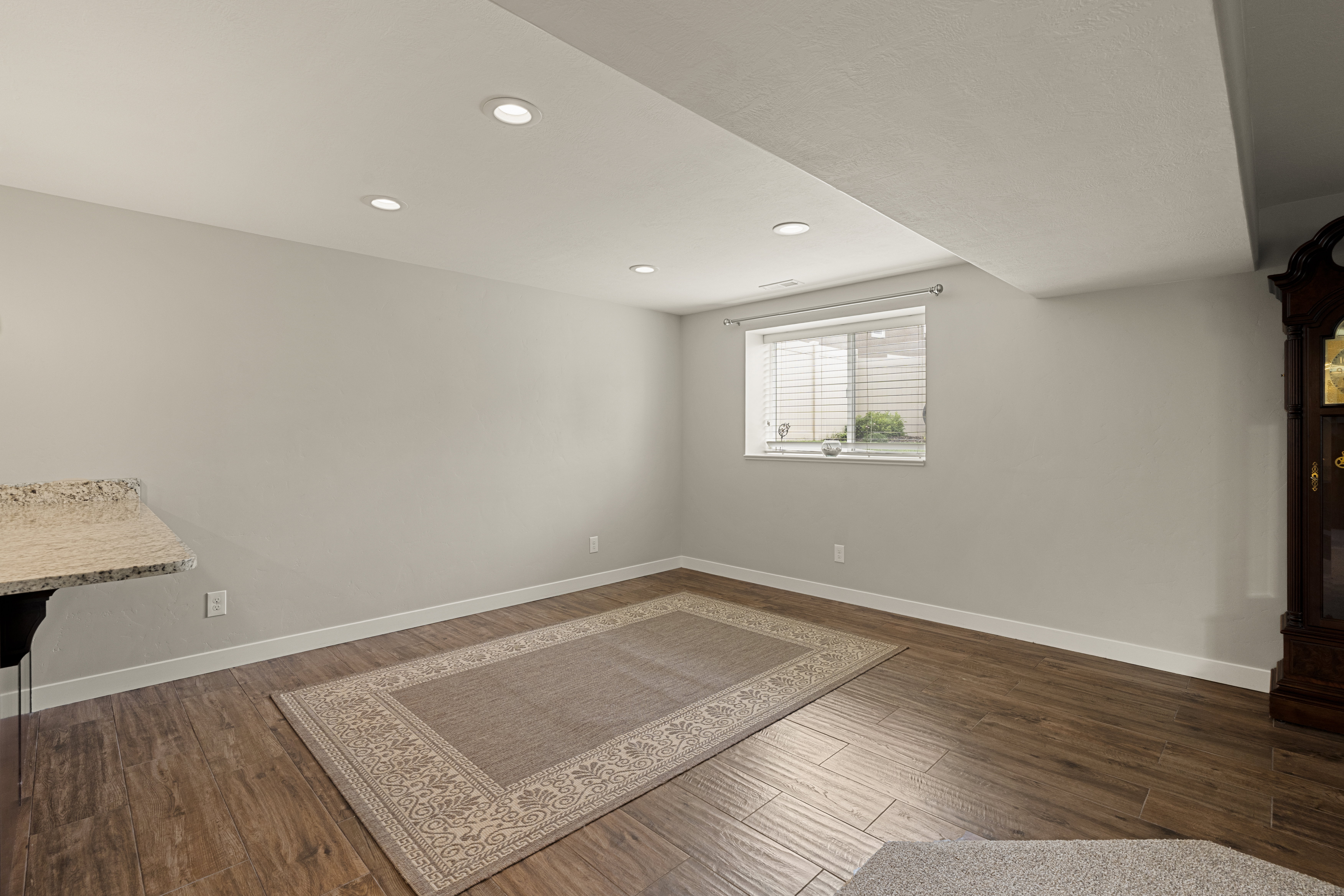 Recessed lighting illuminates this space featuring wood-finish flooring, light gray walls, white baseboards, and a window with blinds