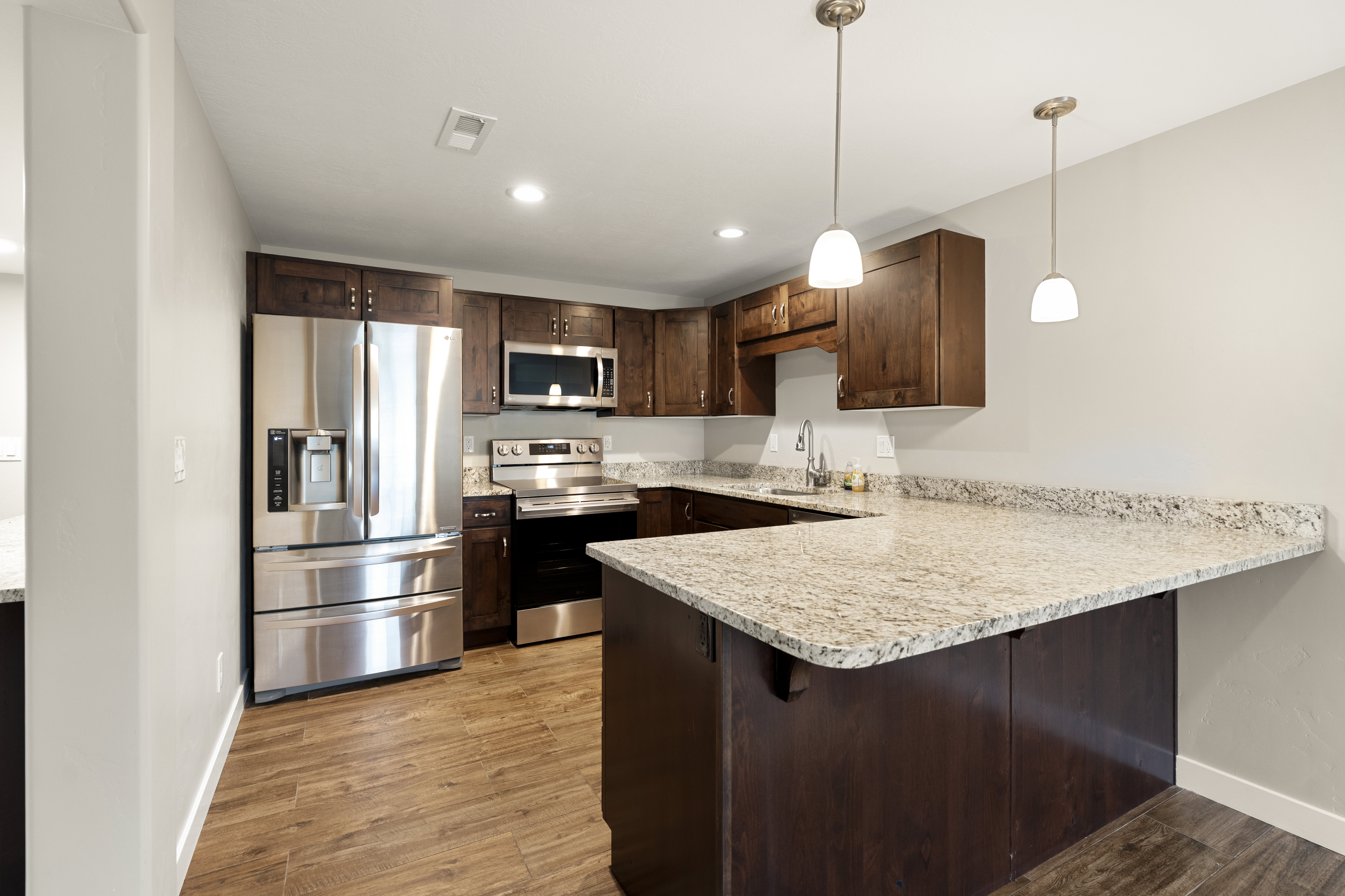 Modern kitchen featuring wood-finish flooring, stainless steel appliances, dark wood cabinetry, granite countertops, and two pendant light fixtures