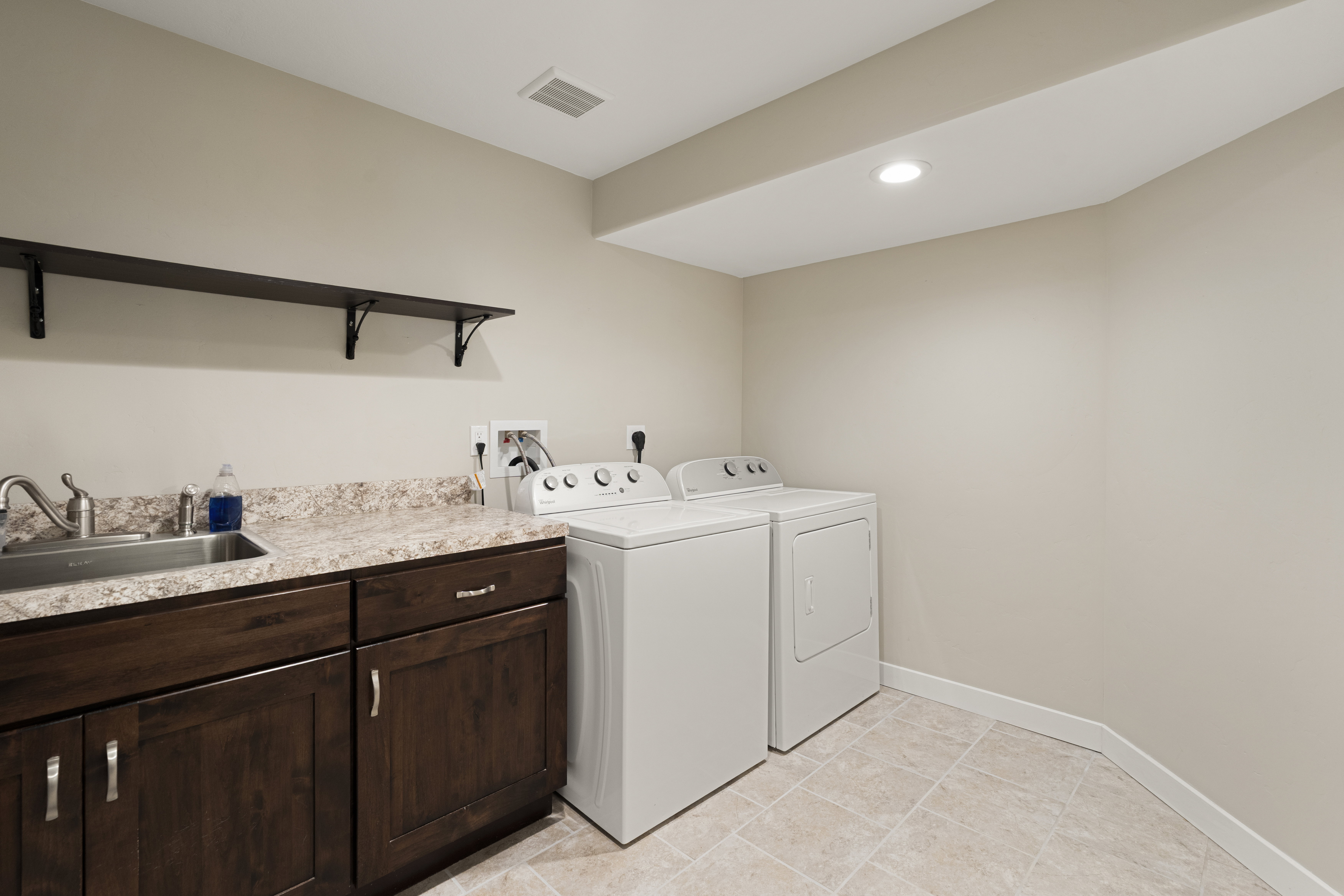 Functional utility room featuring dark wood-finish cabinetry, a stone-look countertop, an integrated sink with a chrome faucet, and tiled flooring