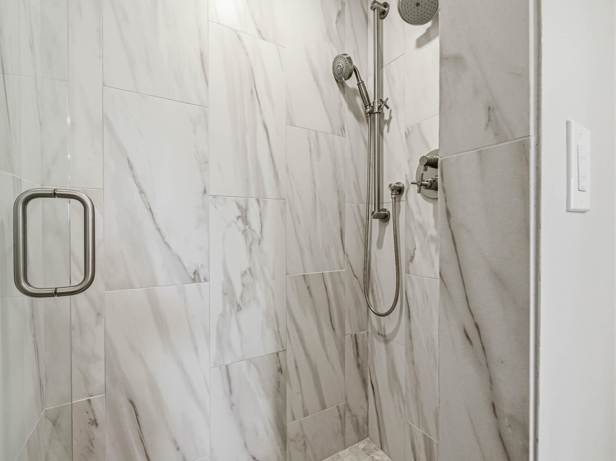 Spacious shower featuring floor-to-ceiling marble-look tile, a frameless glass door with brushed nickel hardware, and a dual shower head system