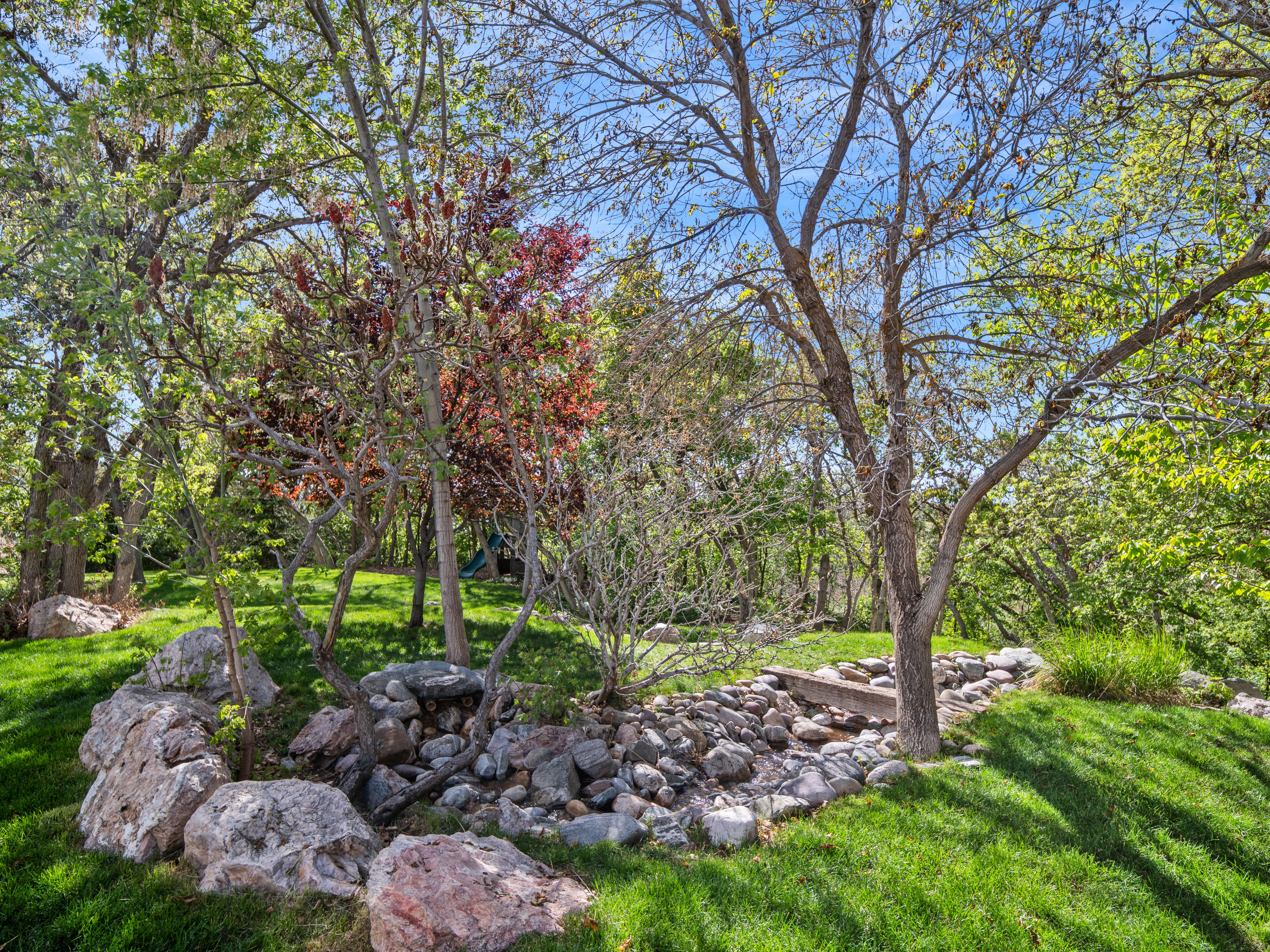 Expansive landscaped grounds featuring mature trees, manicured lawn, natural rock formations, and a decorative dry creek bed with a small wooden bridge element