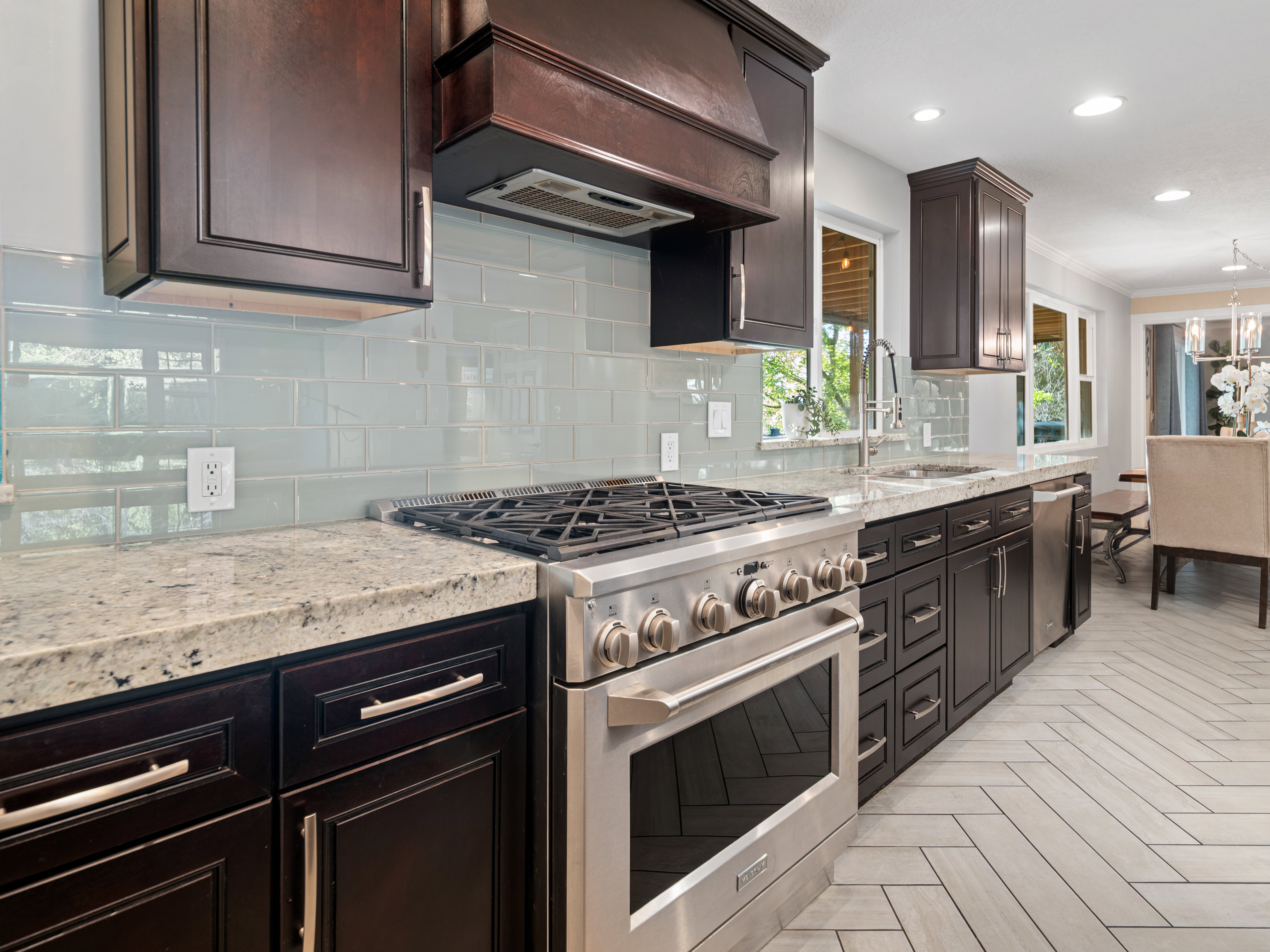 Stainless steel range with a ventilation hood, dark wood cabinetry, light speckled countertops, light blue subway tile backsplash, and light wood-finish herringbone flooring