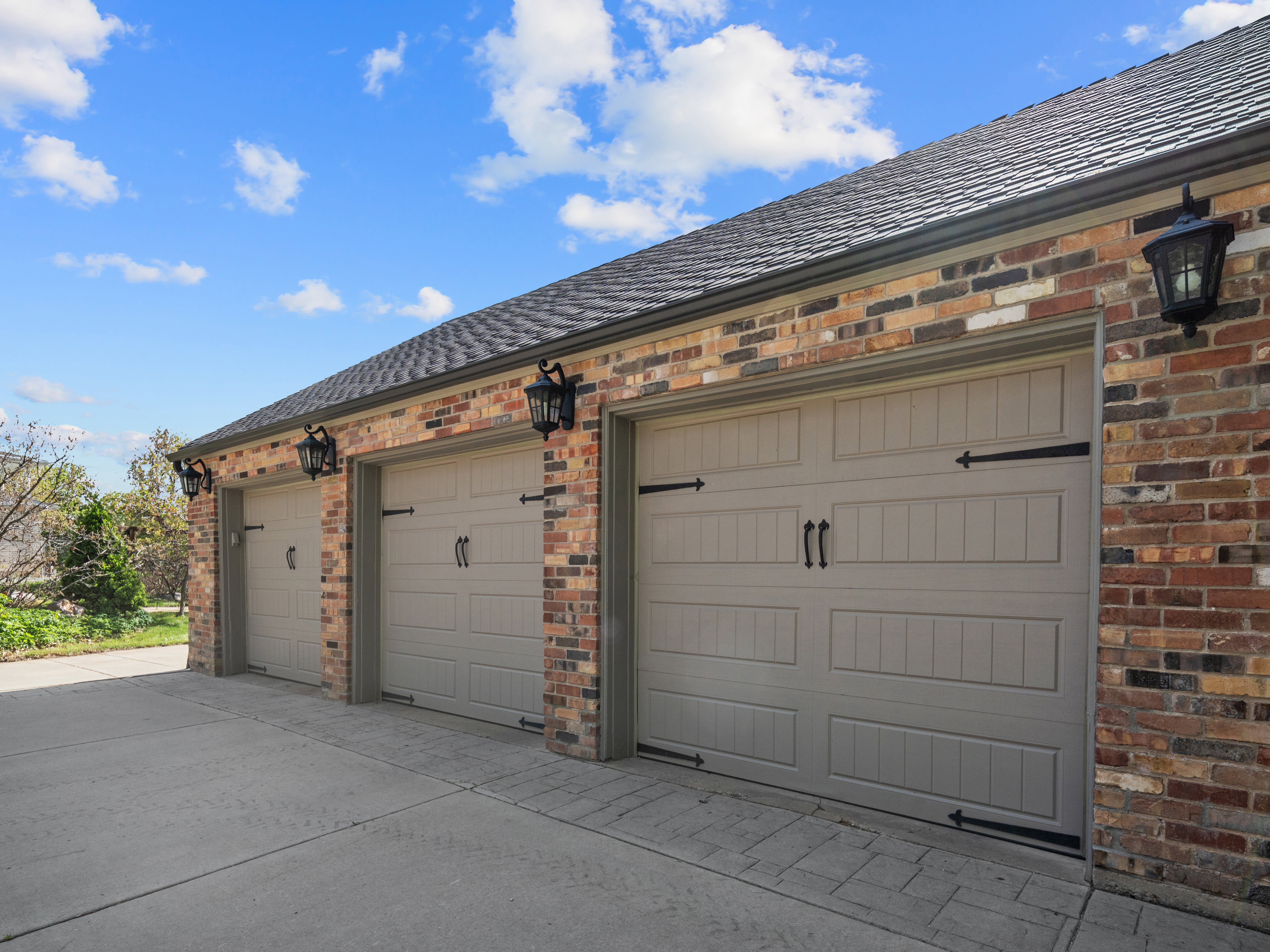 Three-car garage featuring panel-style doors with decorative hardware, brick exterior, exterior wall sconces, and a shingle roof