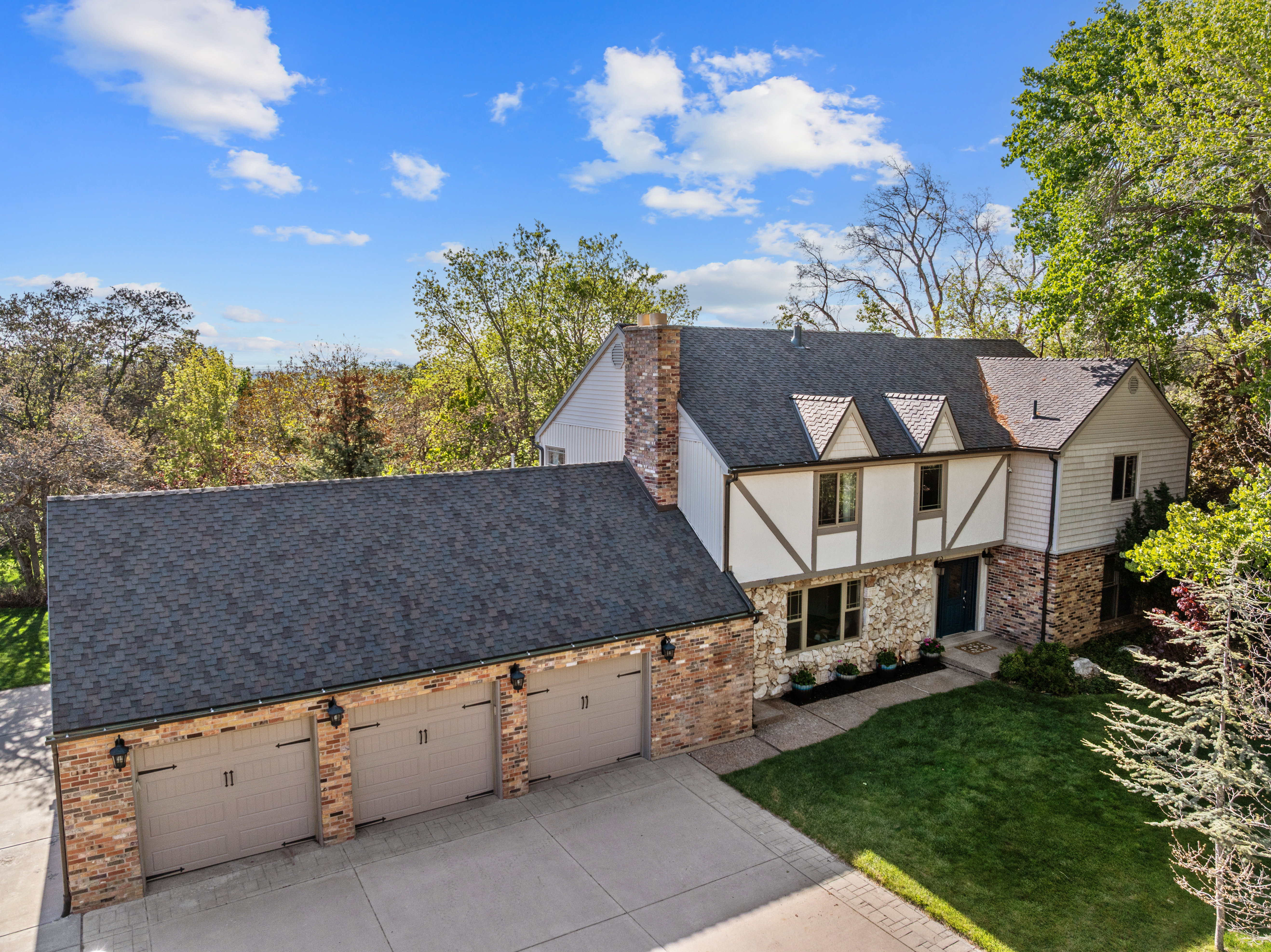 Tudor-style residence featuring a brick and stone facade, three-car garage, multiple gabled dormers, an asphalt shingle roof, and established landscaping