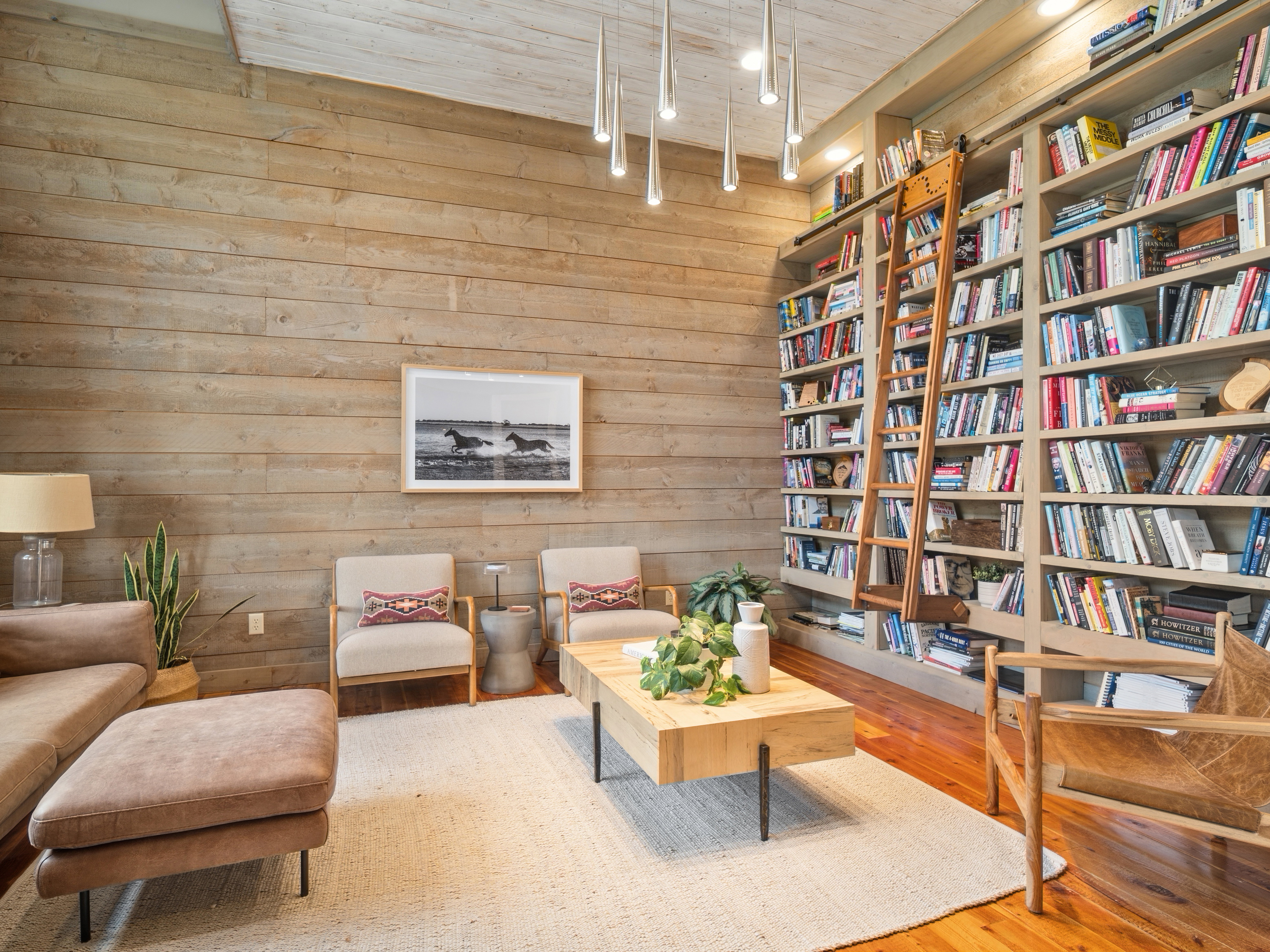 Expansive living space featuring a floor-to-ceiling built-in bookcase with a rolling ladder, wood-plank accent wall, recessed lighting, and modern pendant light fixtures