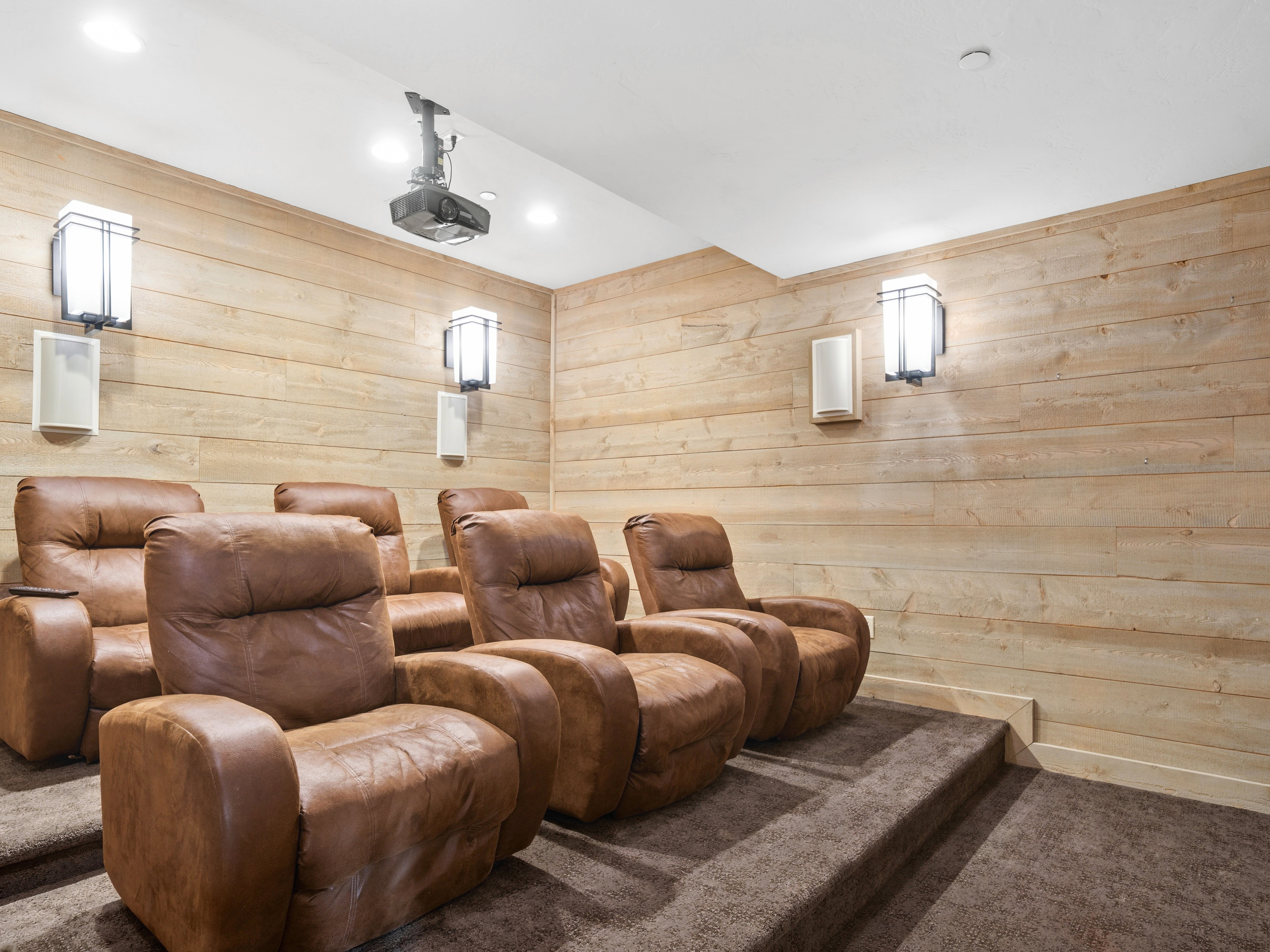 Dedicated media room featuring tiered seating, wood-finish wall paneling, wall-mounted sconces, recessed lighting, and a ceiling-mounted projector