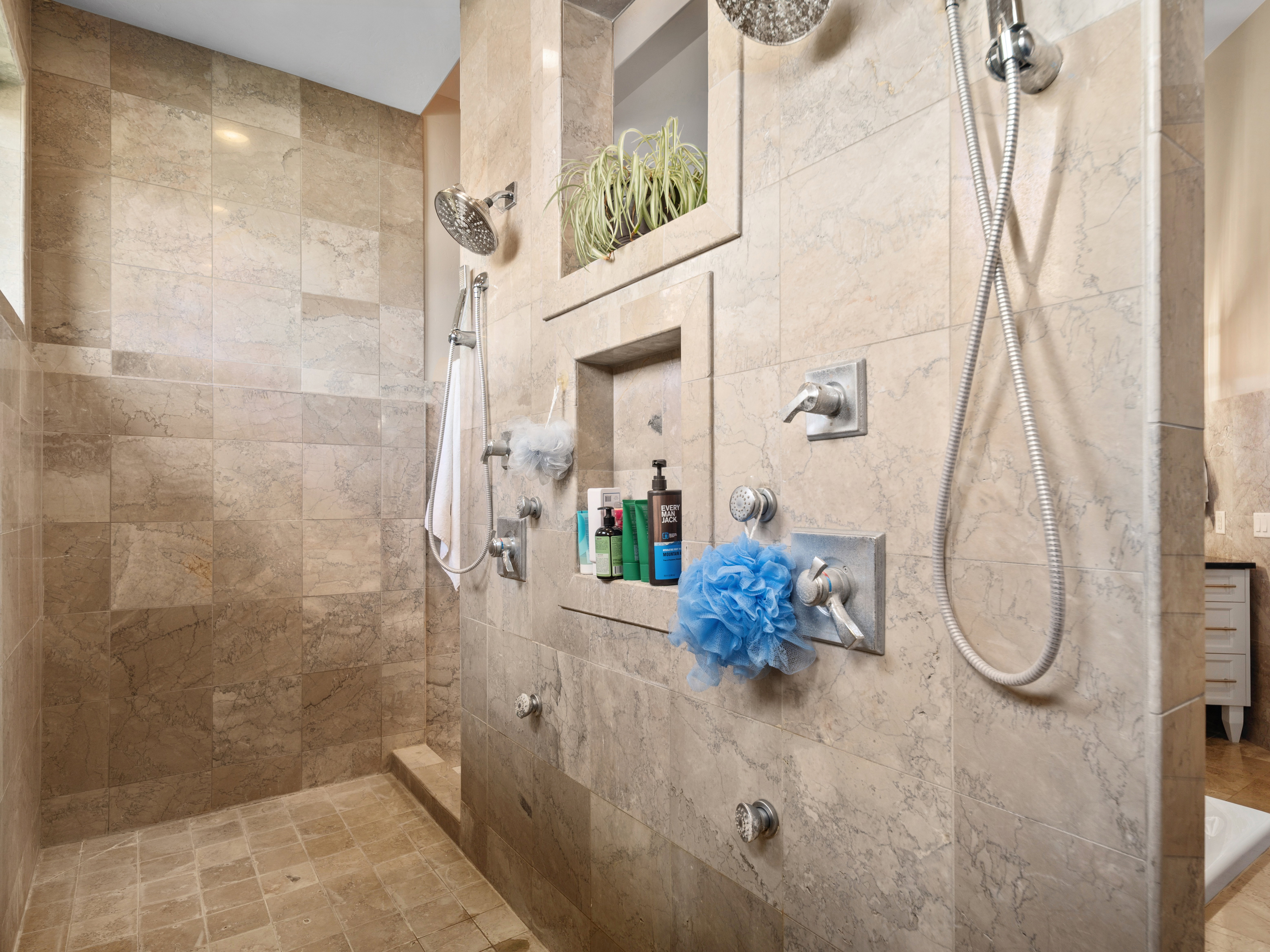 Spacious walk-in shower featuring natural stone-finish wall and floor tiling, multiple shower heads, and built-in shelving niches