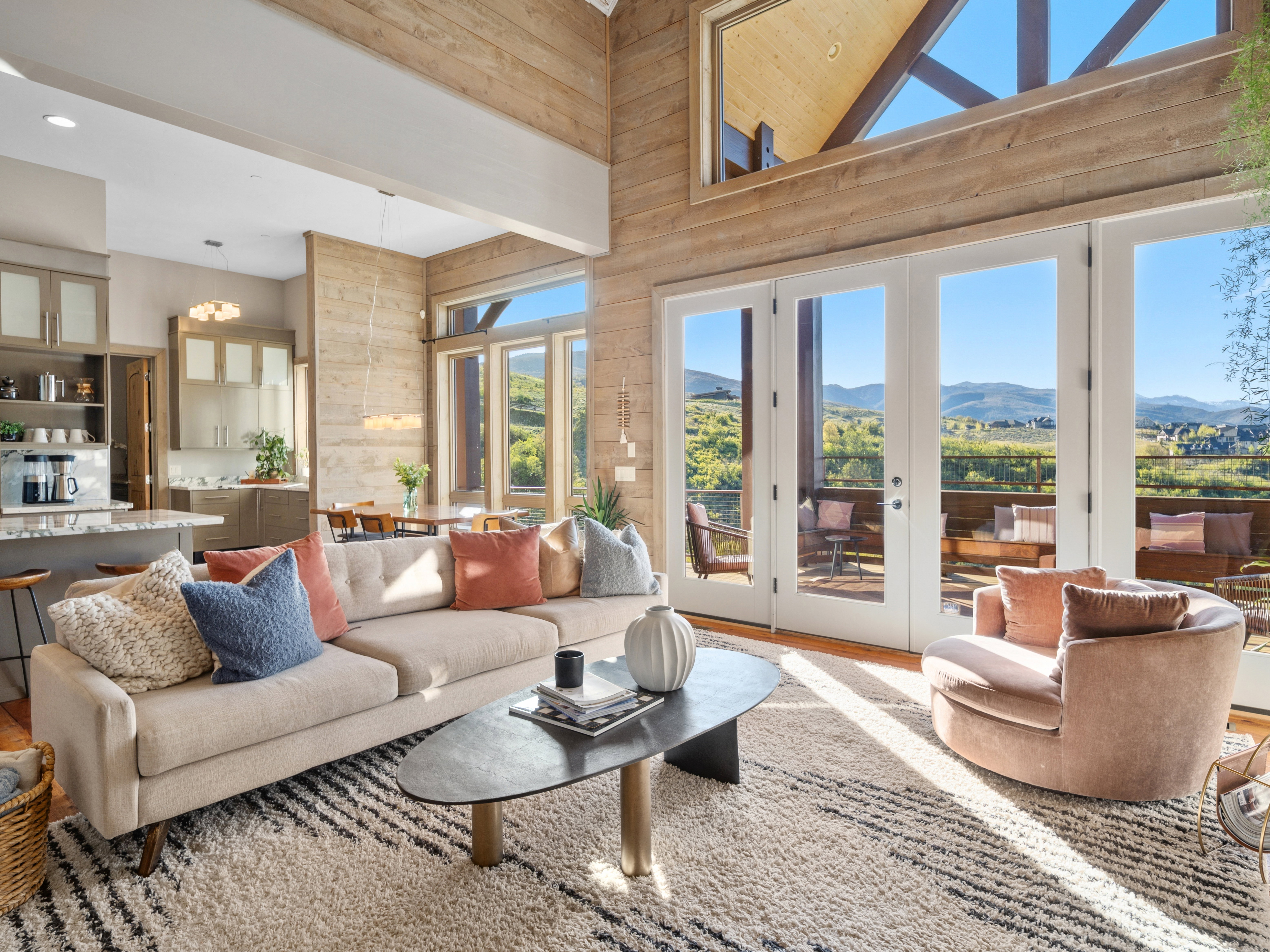 Great room featuring soaring ceilings with exposed wood beams, expansive glass doors opening to an outdoor deck, and a contemporary kitchen with a large island and pendant lighting